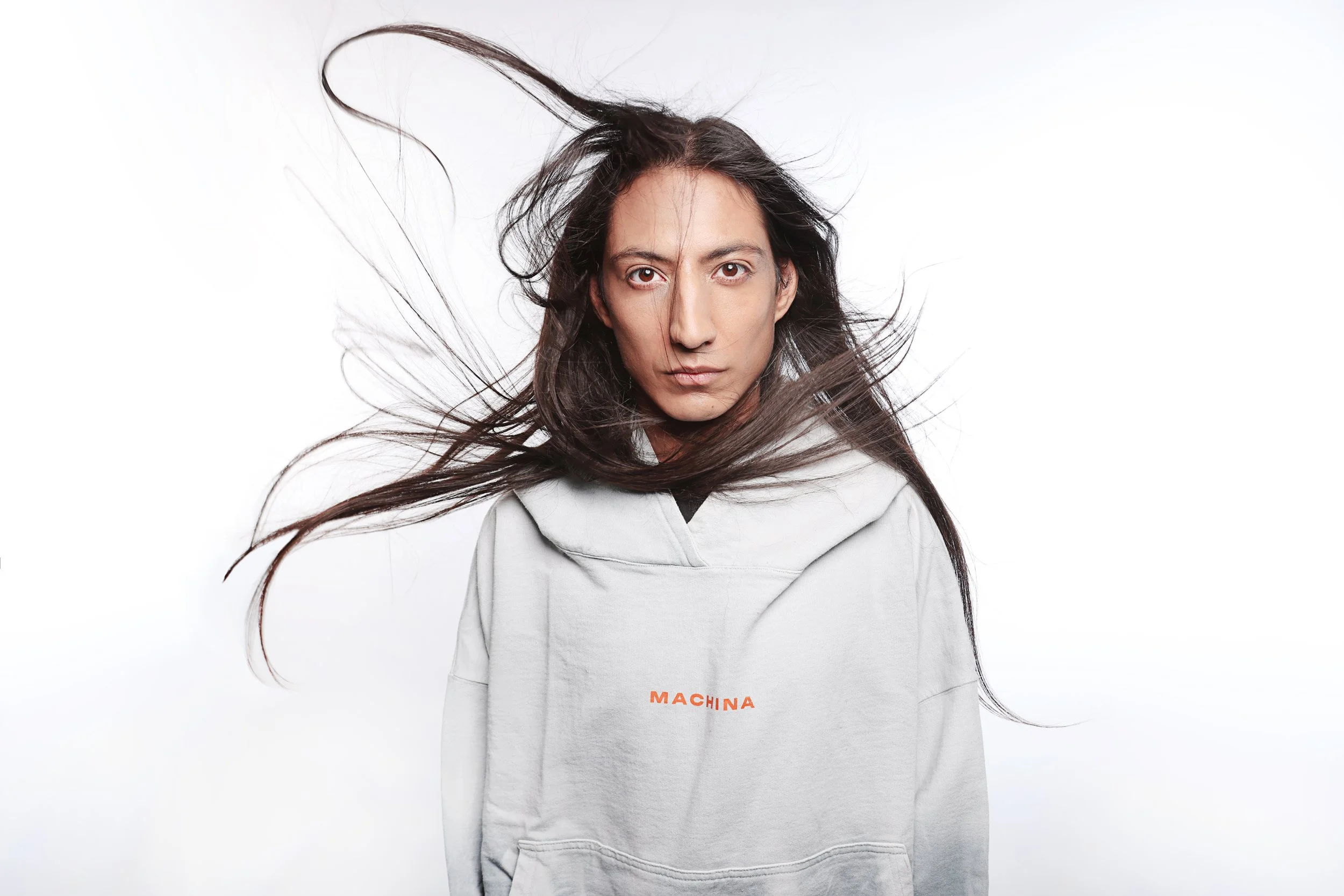 A person with long dark hair and brown eyes wears a gray hoodie with the word 'MACHINA' printed in orange on the chest, standing against a plain white background.