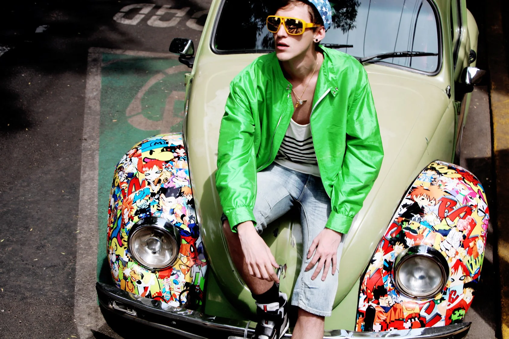 A person sitting on the front of a vintage Volkswagen Beetle with a colorful anime-themed wrap on the hood and fenders. The person is wearing a bright green jacket, striped shirt, light jeans, a blue cap, large yellow sunglasses, and gold jewelry. Th