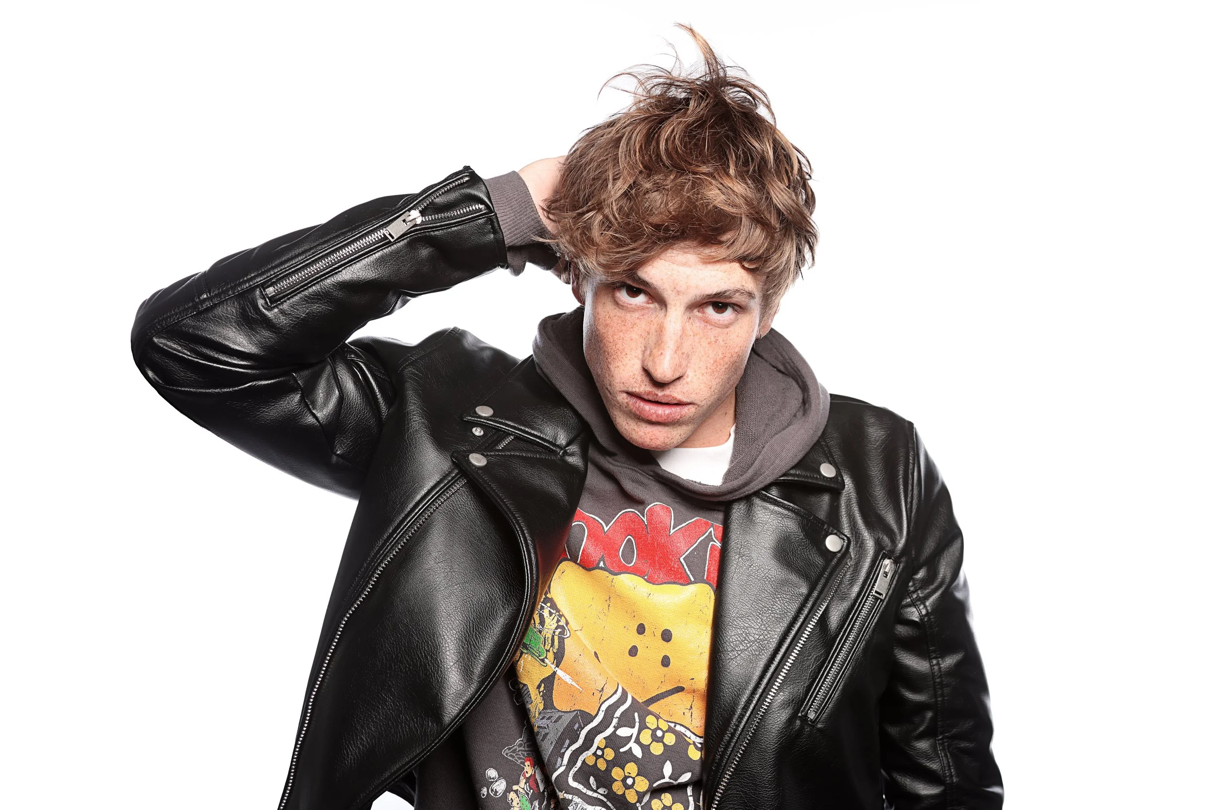 Young man with curly red hair wearing a black leather jacket and a colorful graphic t-shirt, with one hand behind his head, looking into the camera against a white background.
