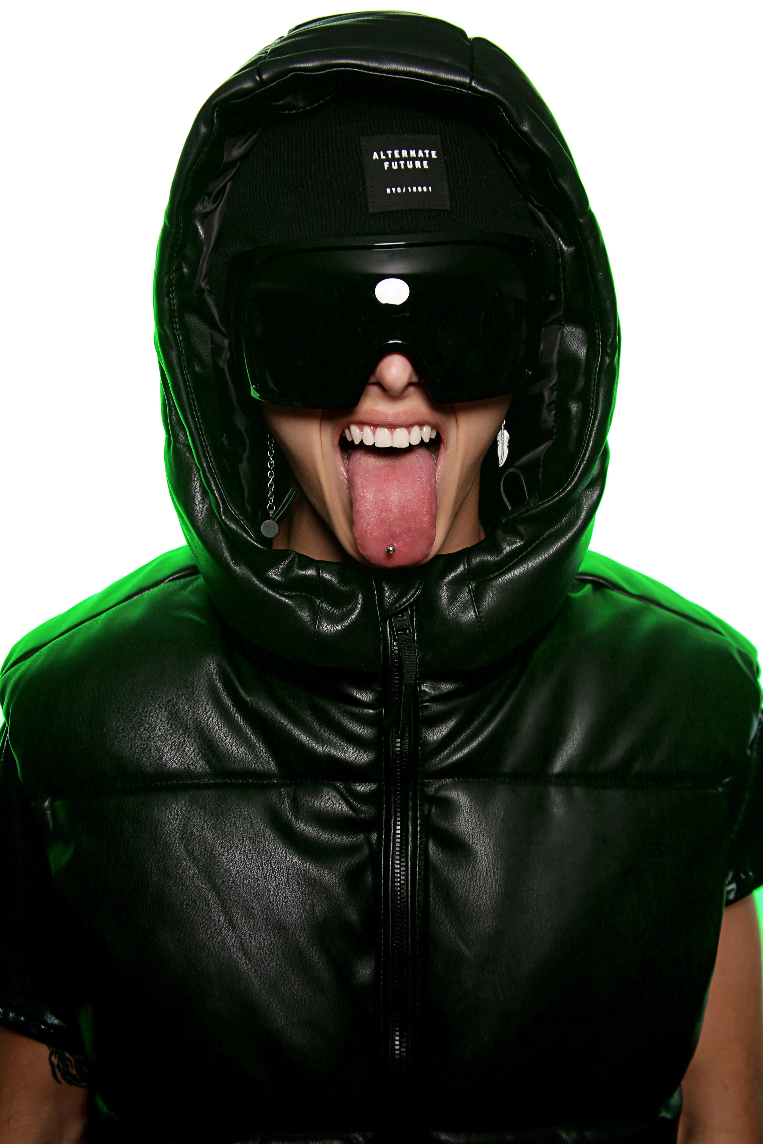 Person wearing a black leather jacket, a helmet with a visor, and a black patch with white text that says 'ALTERNATE FUTURE NYC/1001', sticking out their tongue with a piercing, and showing a silver leaf-shaped earring.
