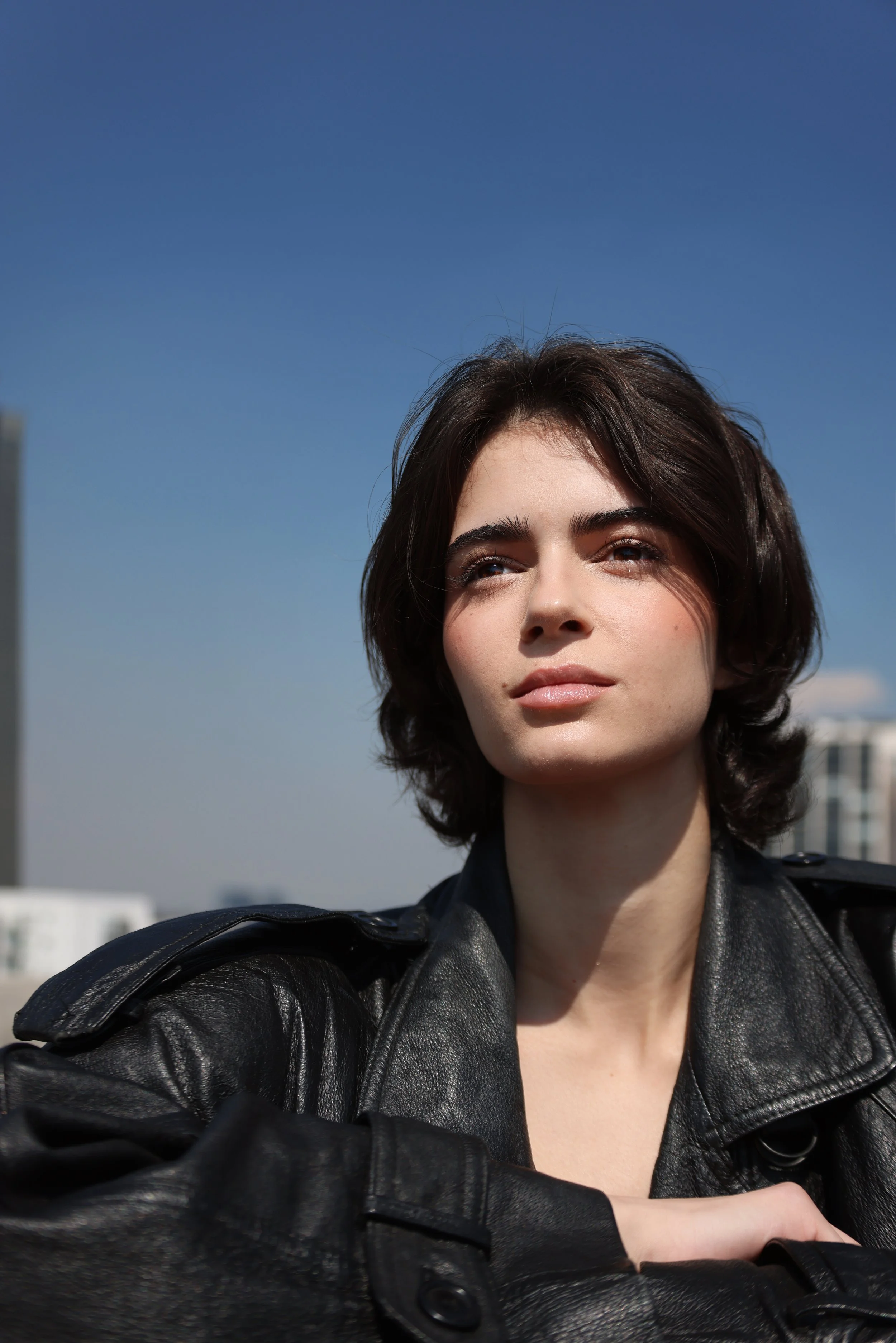 A woman with short dark hair and fair skin wearing a black leather jacket, outdoors against a clear blue sky.