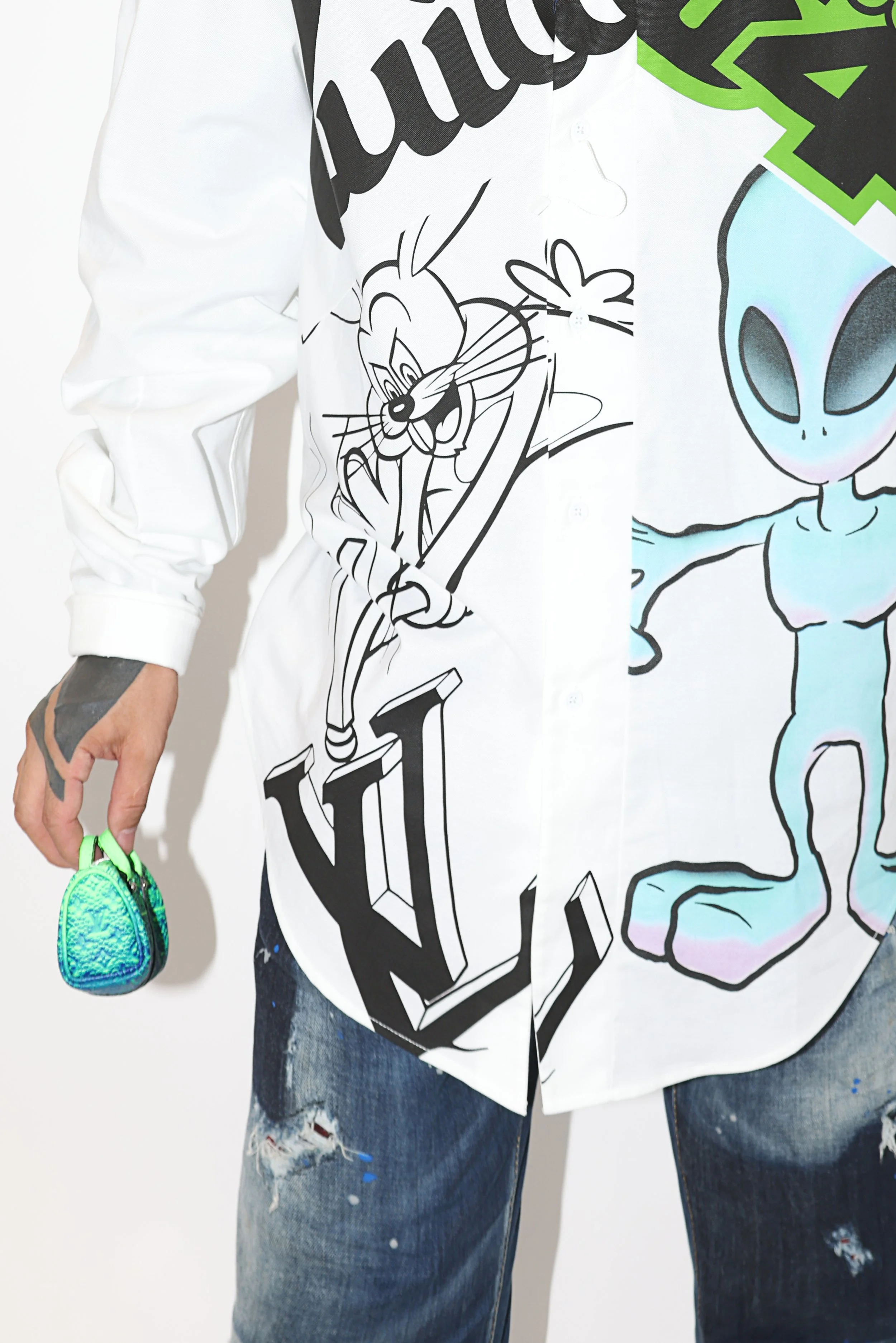 Person wearing a white shirt with black and colorful cartoon characters and text, distressed jeans, and holding a small, shiny green and blue bag in their left hand.