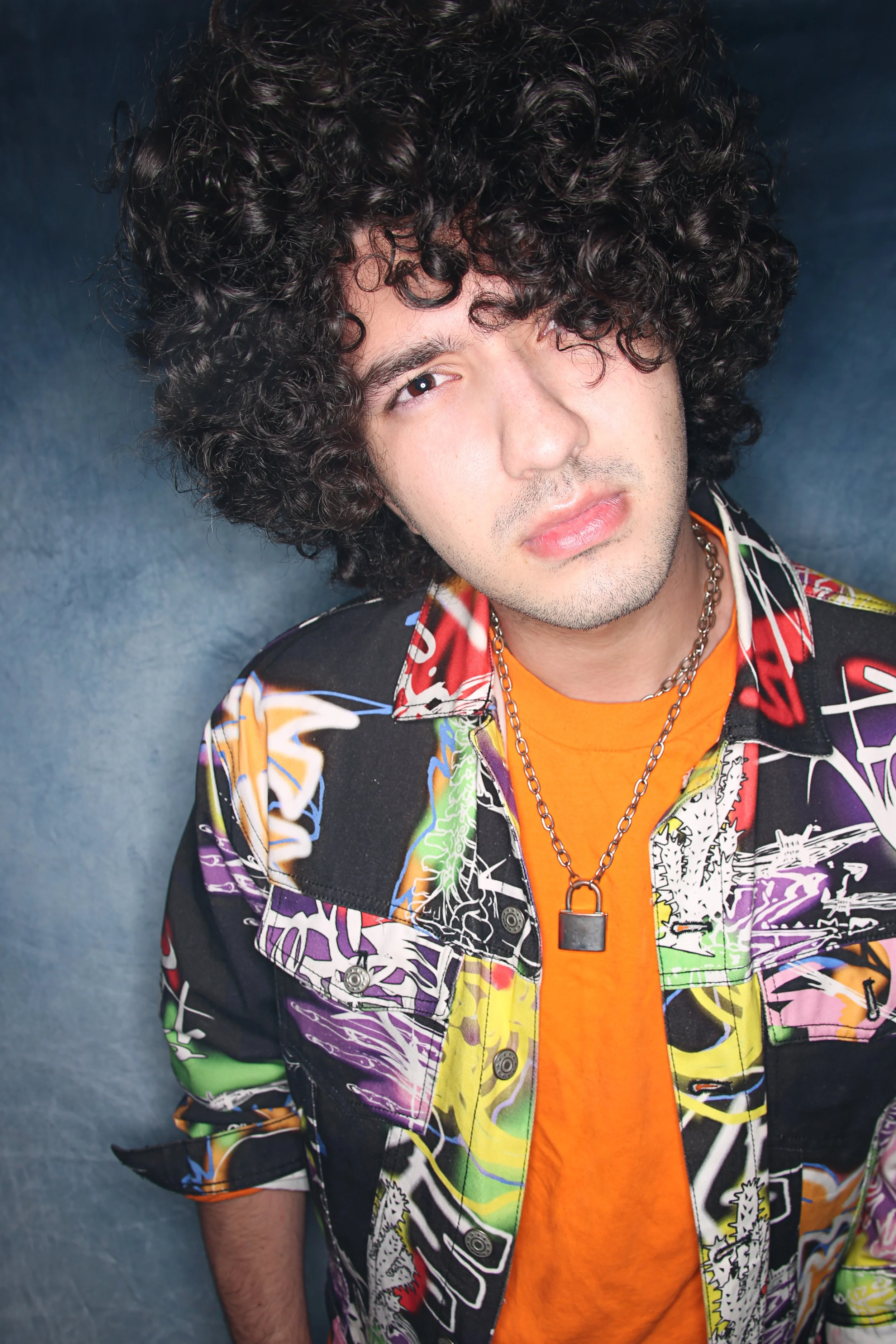 A young man with curly dark hair, wearing a colorful patterned jacket over an orange shirt, and a silver chain with a padlock pendant, stands against a plain grayish-blue background.