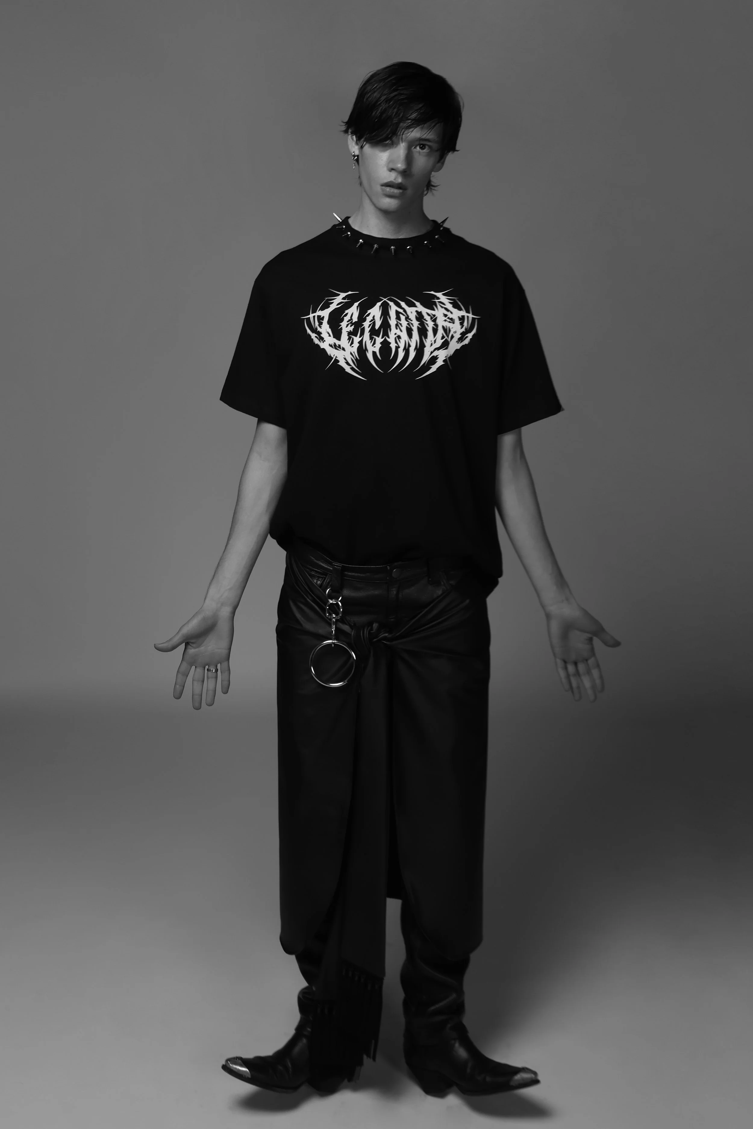 Black and white photo of a person with short dark hair wearing a band t-shirt, wide-legged pants, and boots, standing with arms outstretched against a plain background.