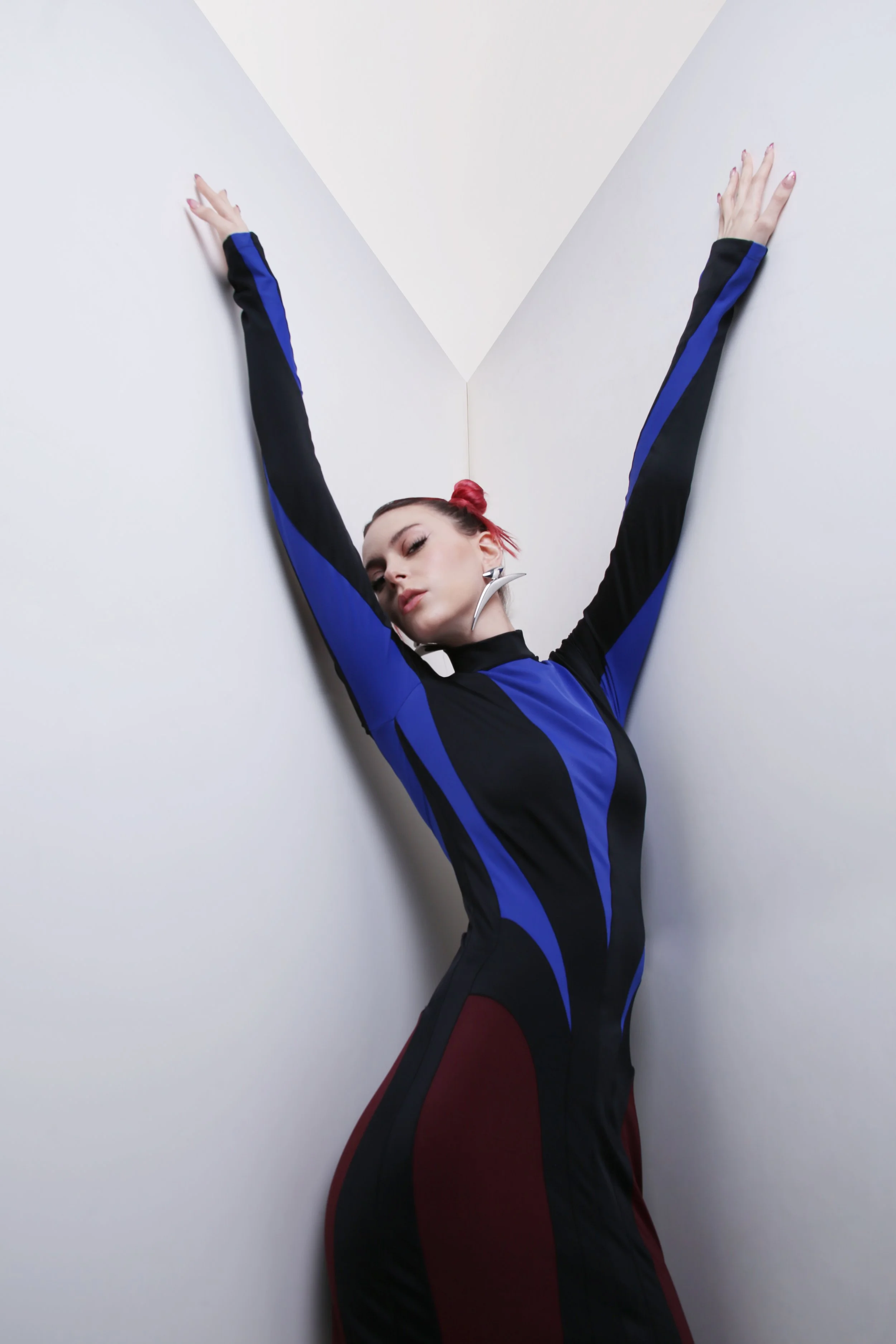 A woman with pink hair styled in a bun, wearing a black and blue athletic outfit, posing with her arms extended upward against a plain white wall.