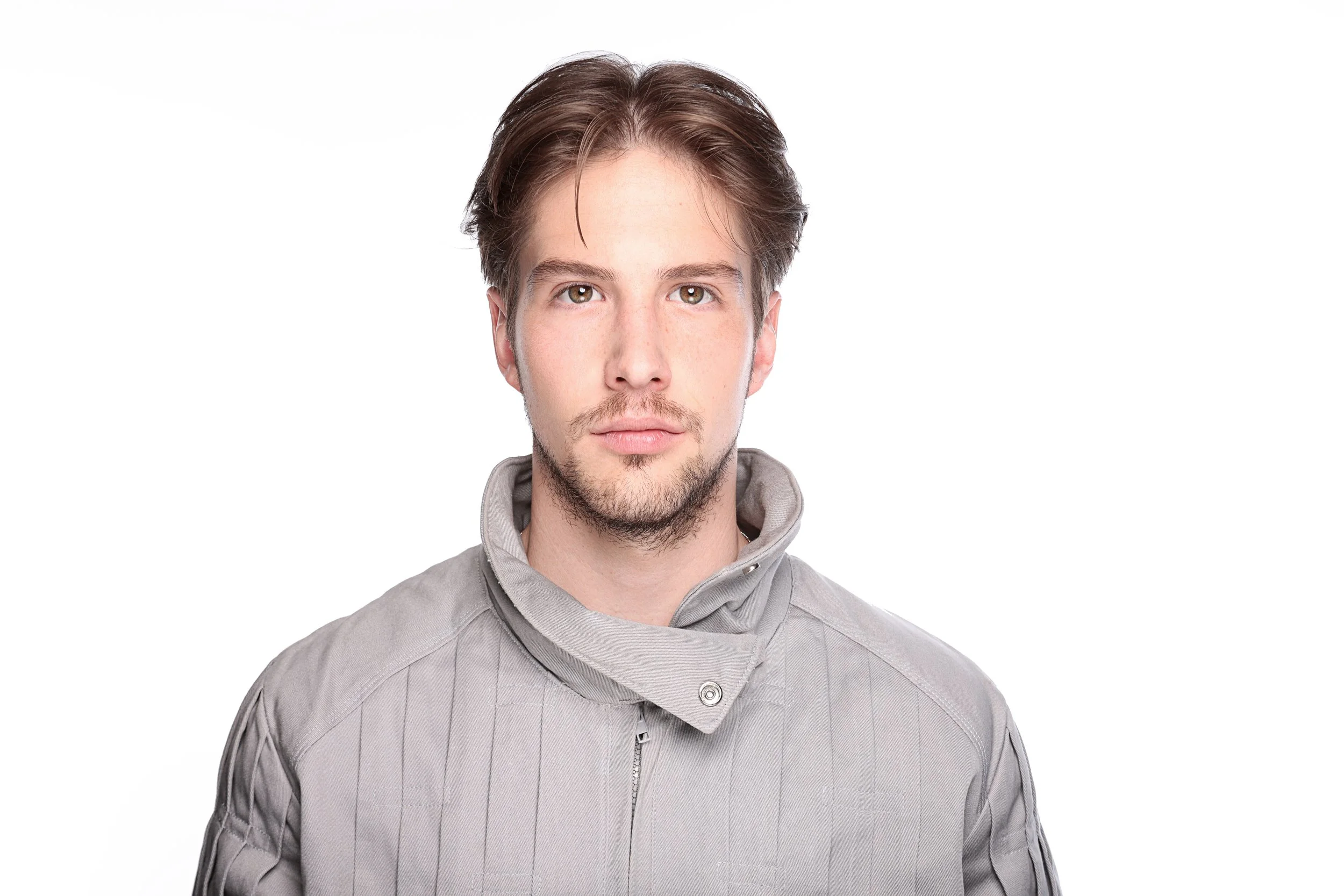 A young man with brown hair and hazel eyes, wearing a light gray jacket with a high collar, standing in front of a plain white background.