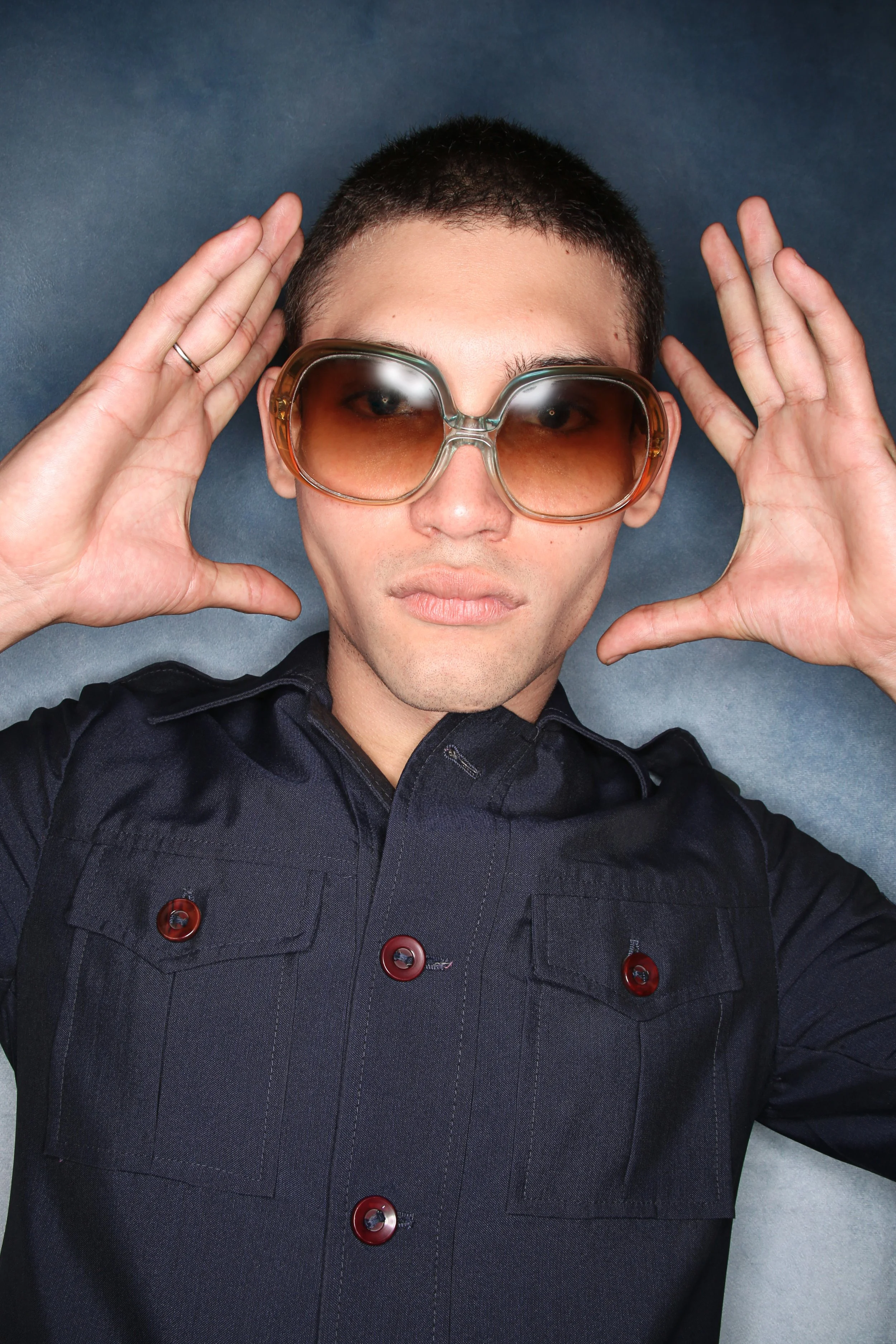 A man with short black hair wearing large brown sunglasses, a dark jacket with red buttons, and making a pose with his hands near his temples against a dark background.