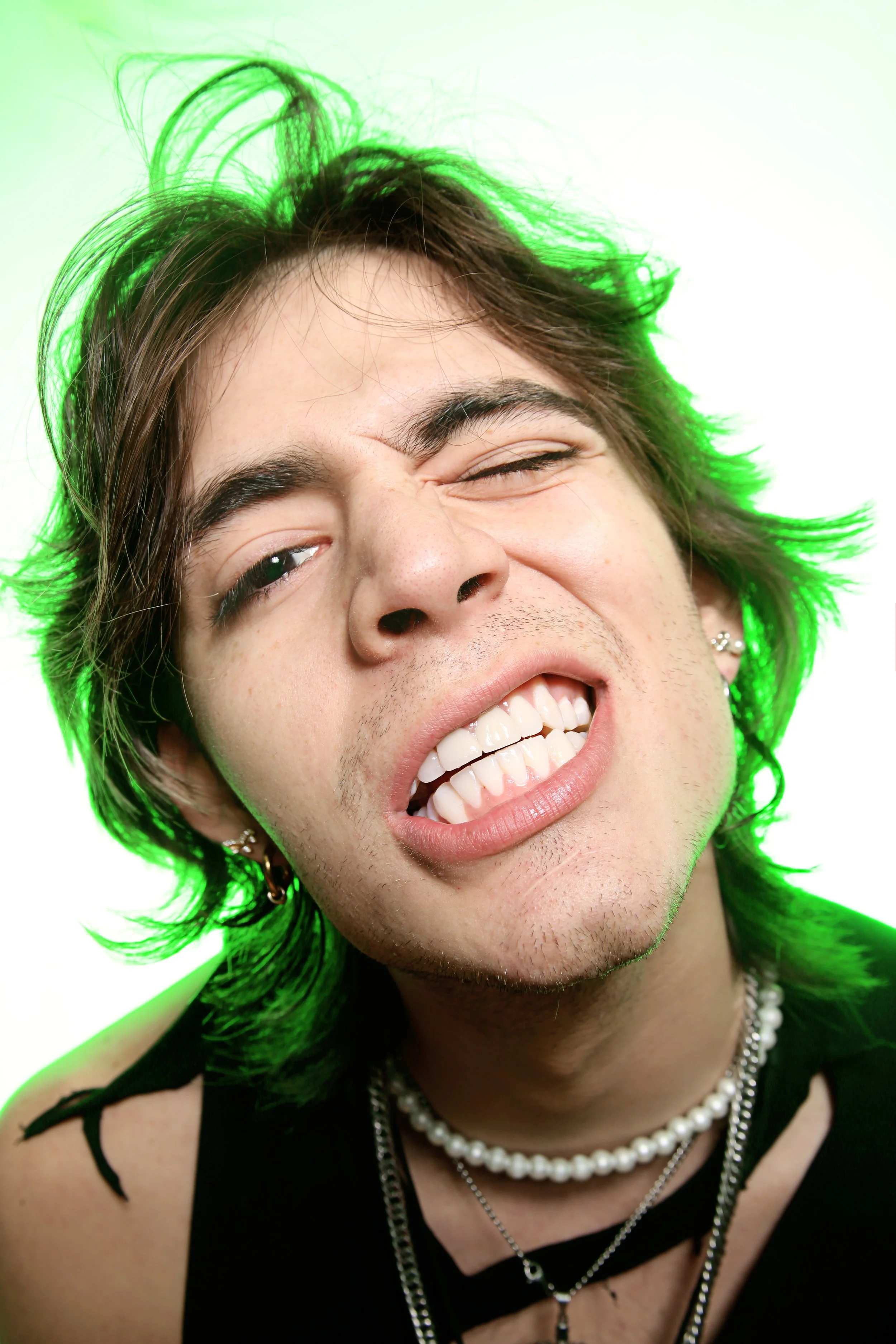 Close-up of a smiling young man with brown hair, some green hair highlights, wearing layered necklaces and earrings, against a white background.