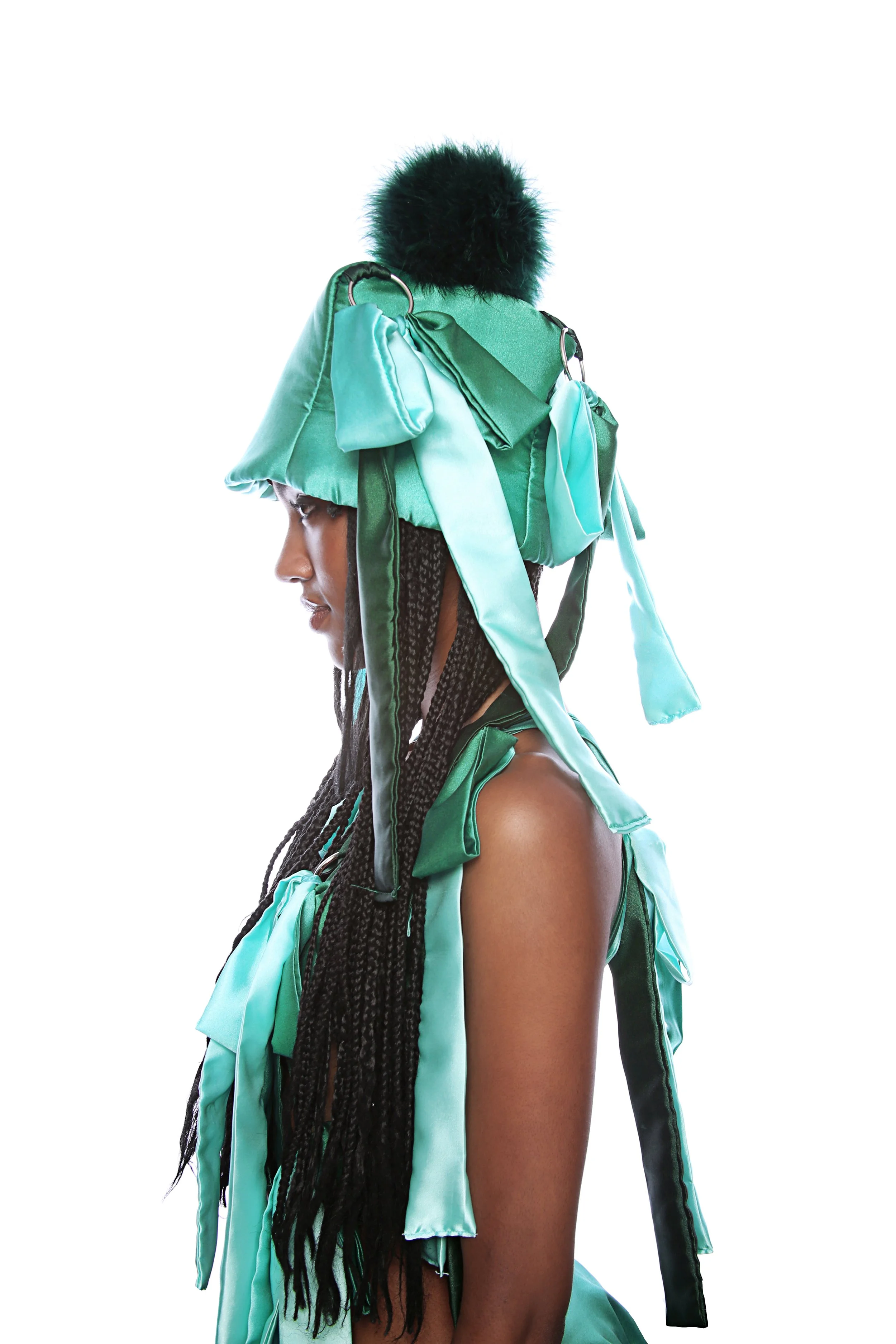 A woman with long braided hair seen in profile view, dressed in flowing green and teal garments, wearing a large matching headpiece with a black fur pom-pom on top, against a plain white background.