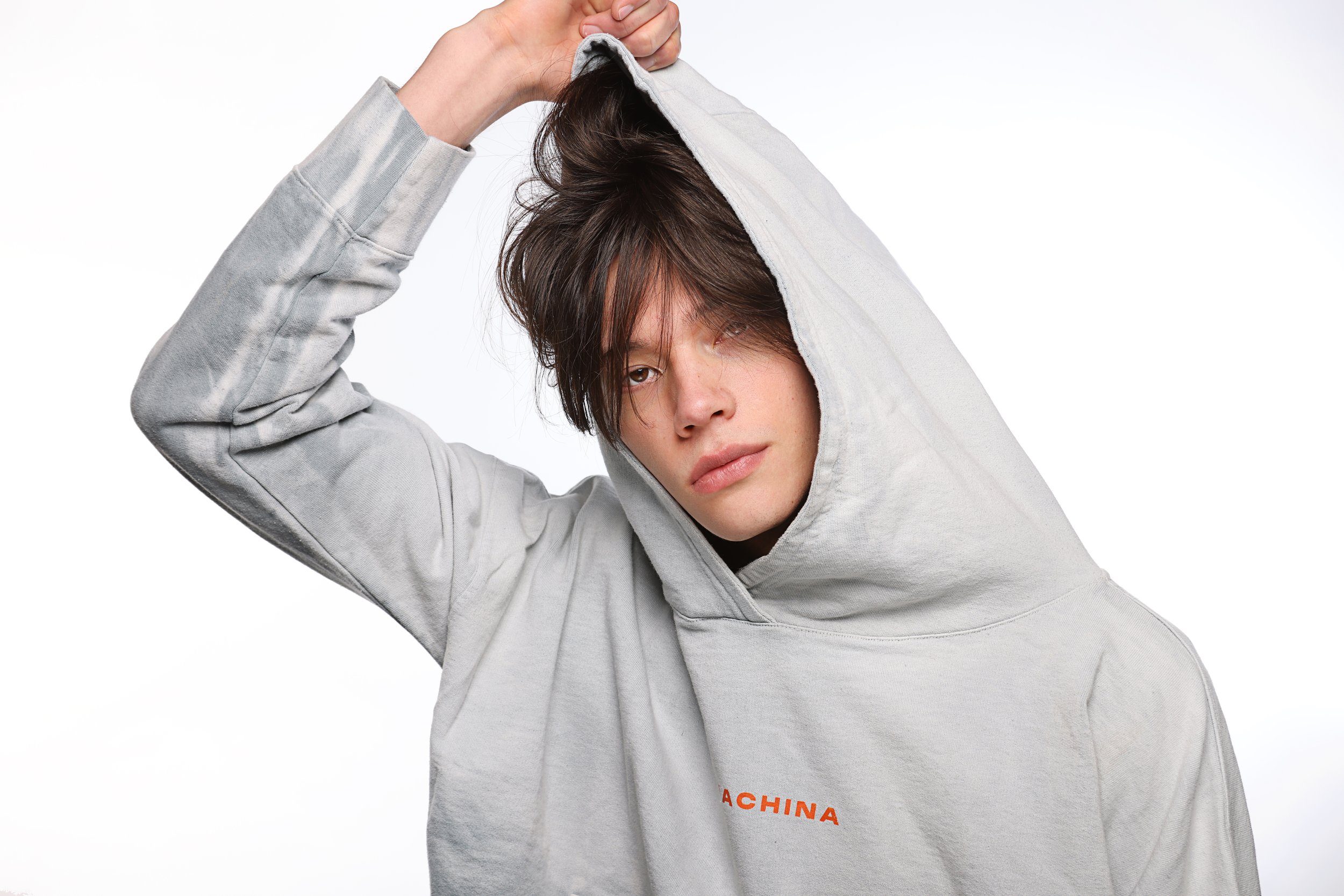 Young person wearing a light gray hoodie with the hood over their head, looking at the camera, against a plain white background.