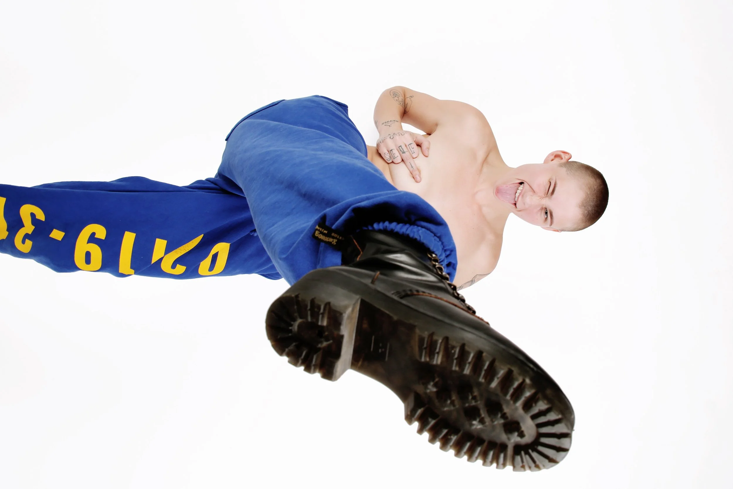 A person with a shaved head and tattoos, wearing a blue jacket and black boots, looking down at the camera while sticking out their tongue, against a white background.