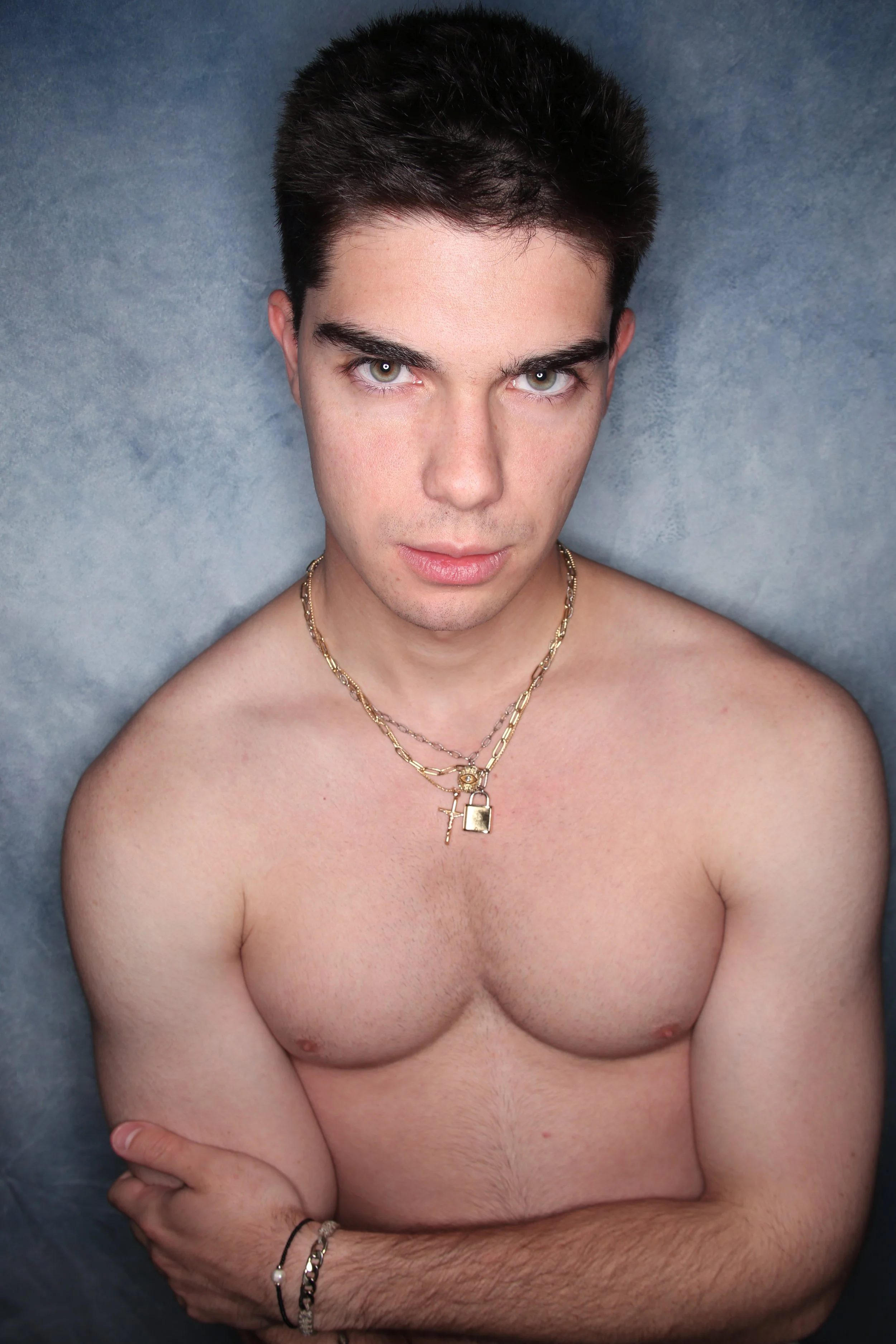 A shirtless young man with dark hair, green eyes, and light skin, crossing his arms, wearing multiple gold necklaces and bracelets, against a textured gray background.