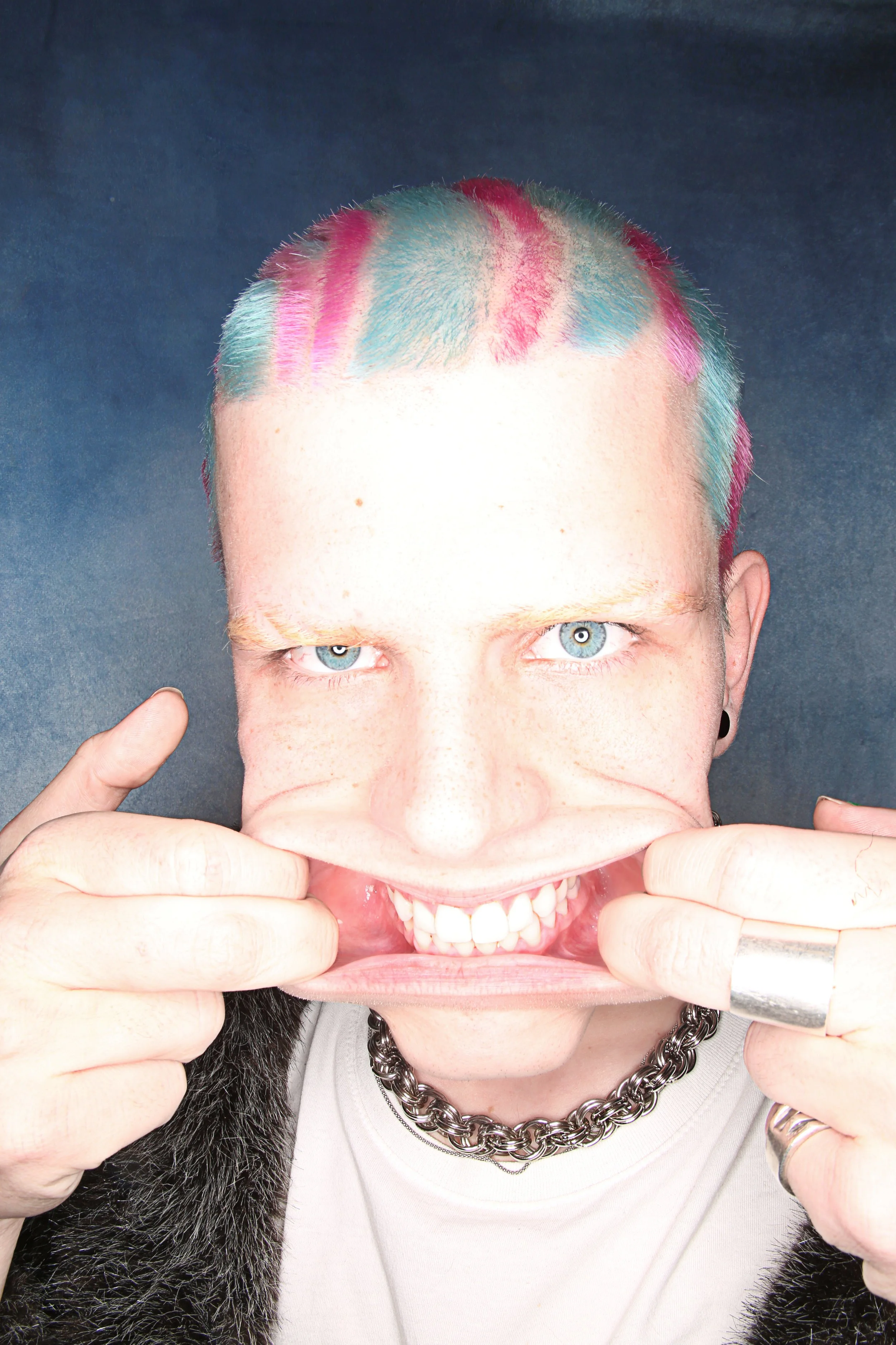 Person with colorful, dyed hair pulling a mask of a smiling face down, revealing real face with bright blue eyes, wearing a silver chain necklace and black fur coat.