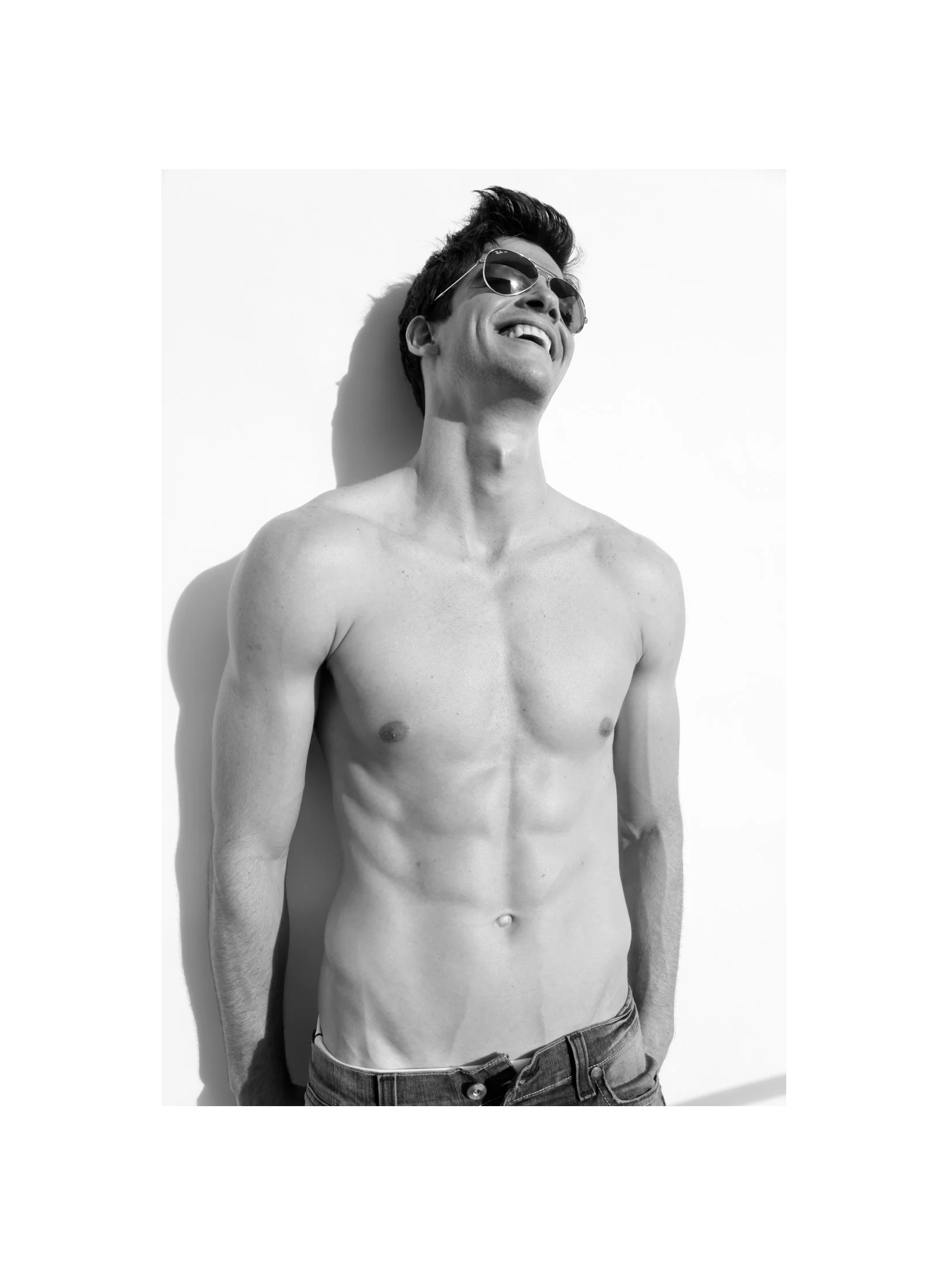 Black and white photo of a smiling young man wearing sunglasses, shirtless, with hands in pockets, facing slightly to the right against a plain wall