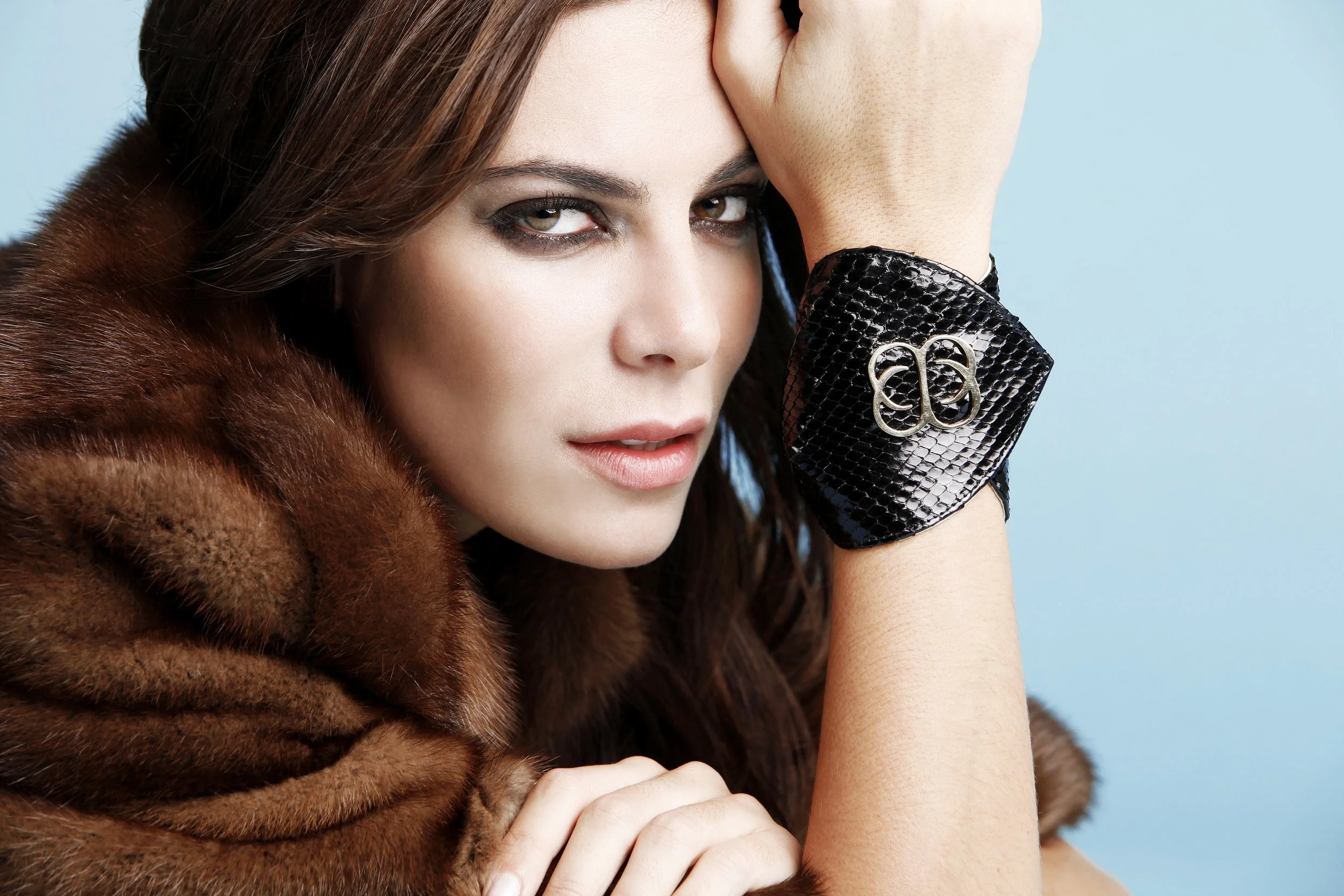 Close-up of a woman with brown hair, hazel eyes, wearing dark eye makeup, a black snakeskin-patterned bracelet with a silver logo, and a brown fur coat against a light blue background.