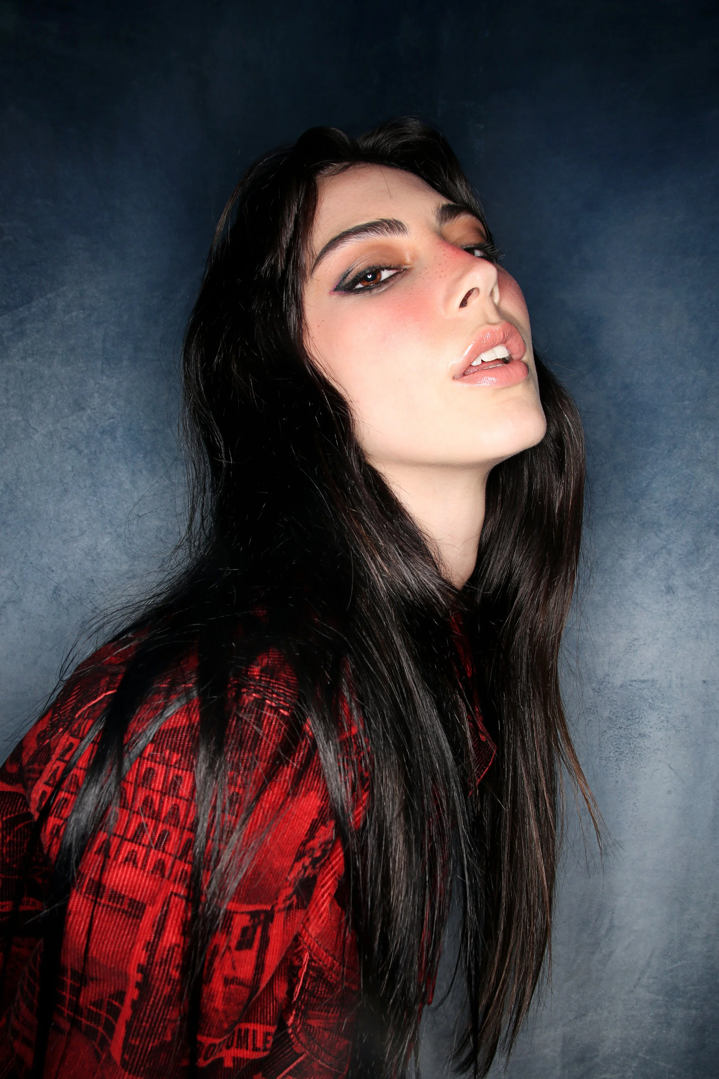Close-up of a woman with long dark hair, makeup, and a red patterned shirt posing against a dark background.