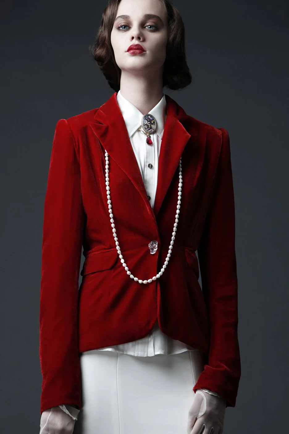 A woman with fair skin, dark brown hair, and red lipstick, wearing a red velvet blazer, white gloves, a white shirt with black buttons, a pearl necklace with a skull-shaped pendant, and a brooch with a skull design.