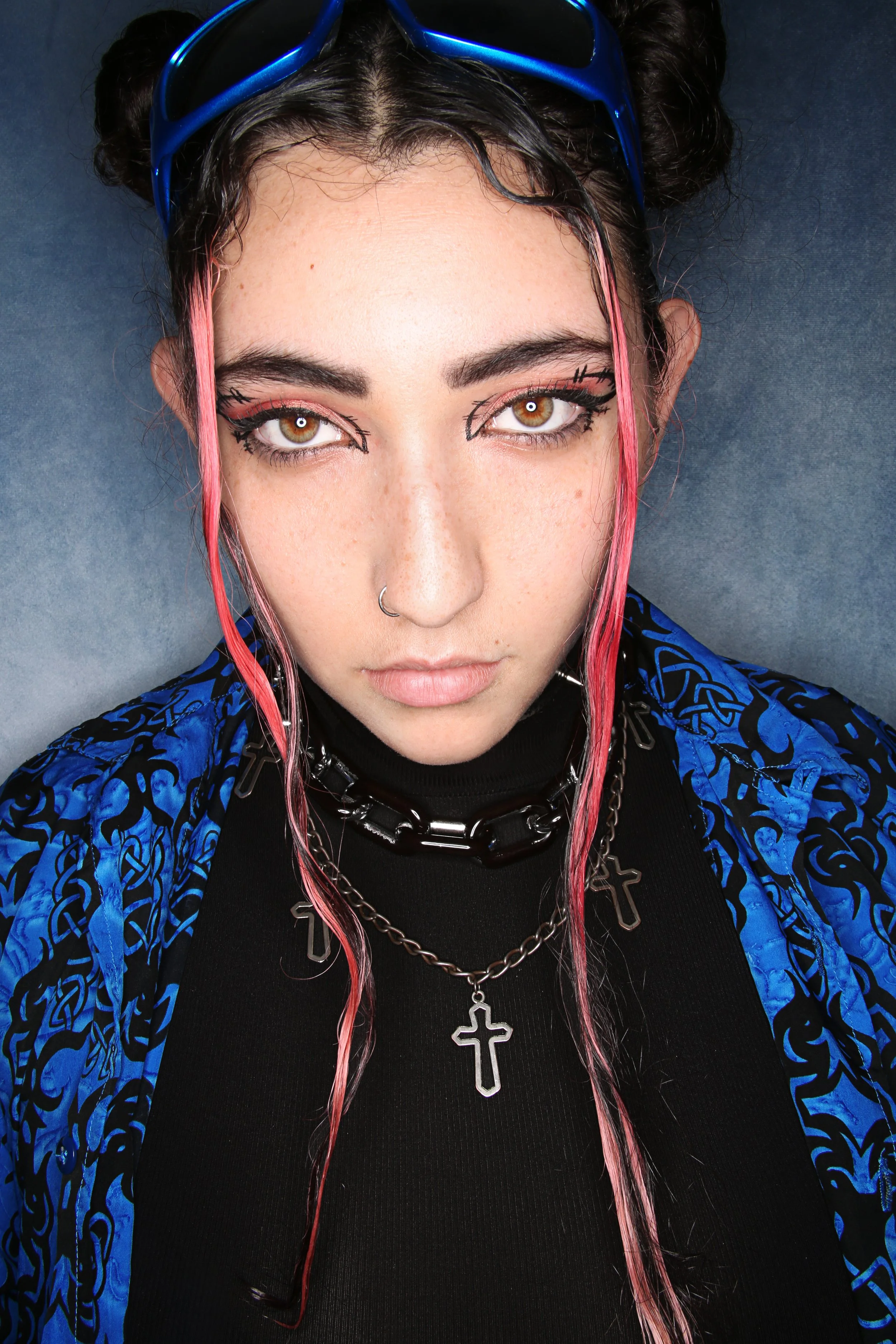 Close-up of a young woman with brown eyes, dark hair with pink highlights, wearing sunglasses on her head, black eyeliner, layered necklaces including a cross, and a blue patterned jacket over a black top.