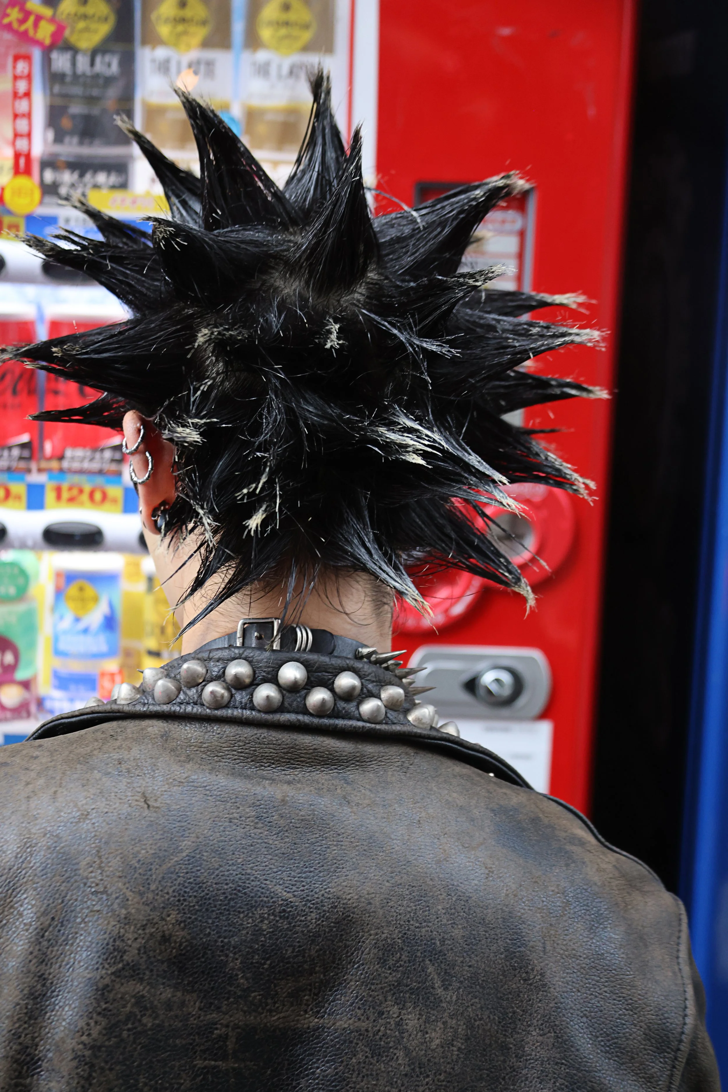 Back of a person with spiky black hair, wearing a leather jacket with metal studs, standing in front of a vending machine.