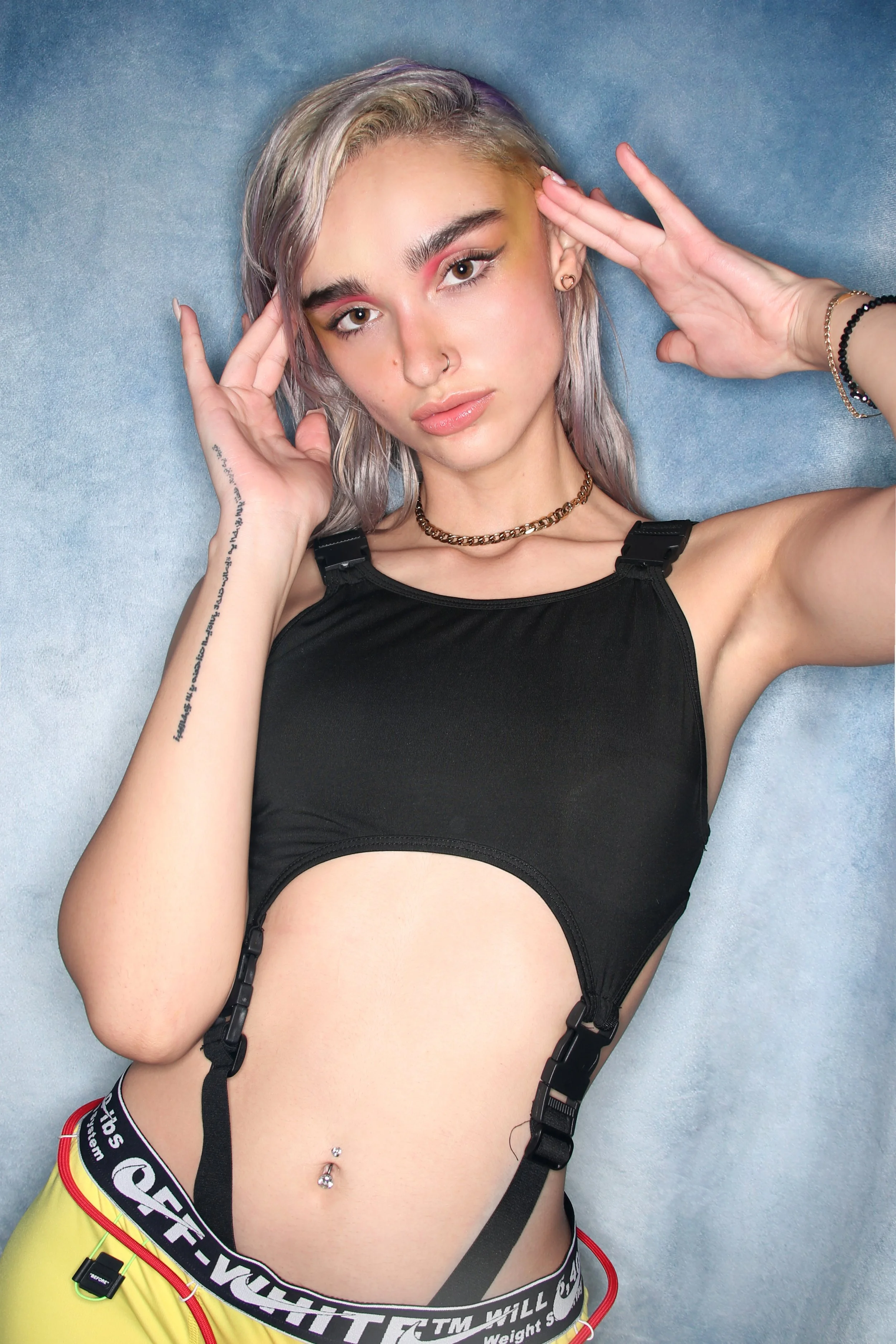 A young woman lying on her back on a blue surface, wearing a black sports top and yellow shorts with a black waistband. She has silver hair, light makeup, a nose ring, a belly button piercing, and several bracelets on her wrists. Her hands are placed