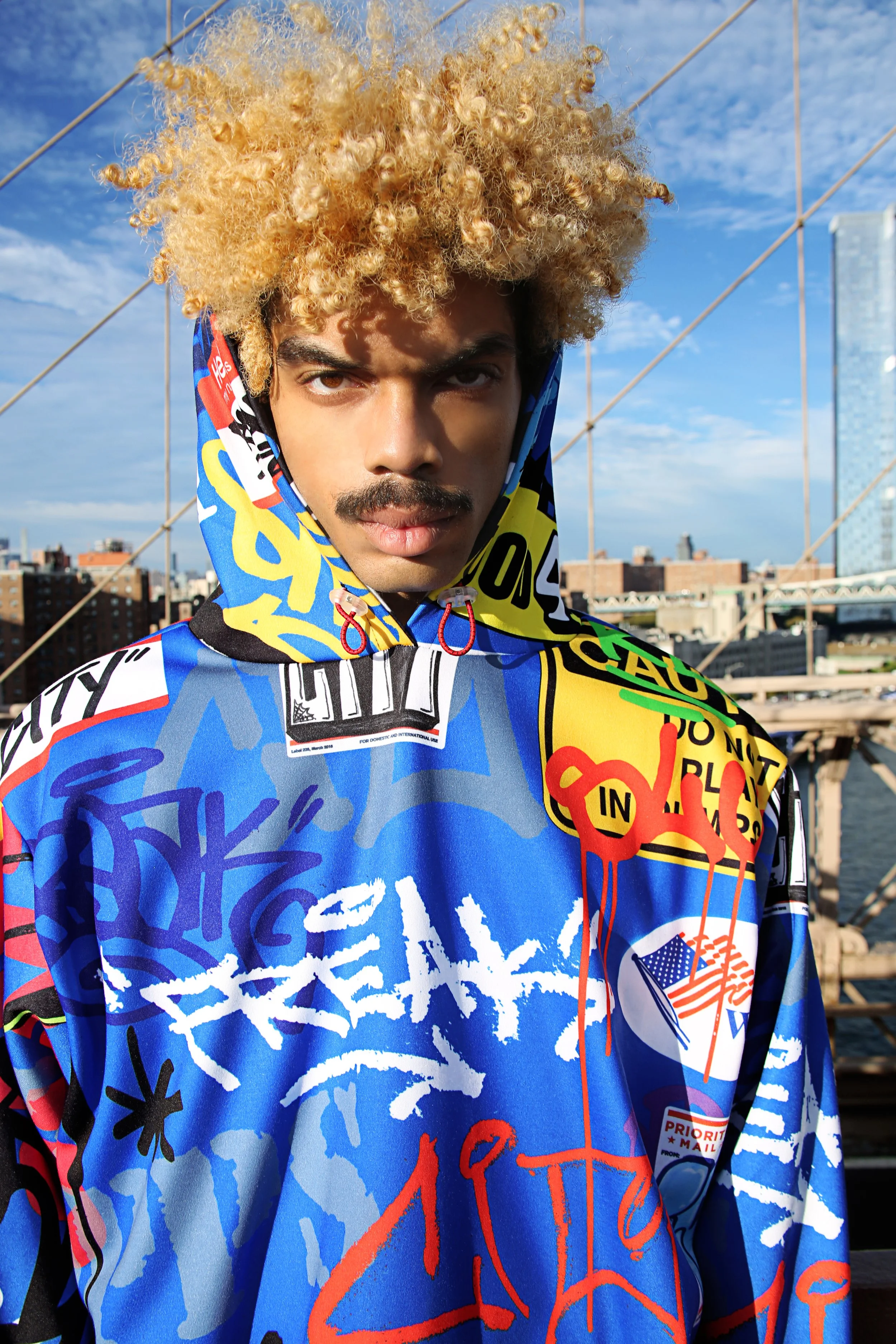 A young man with a blonde, curly hairstyle wearing a colorful, graffiti-style jacket standing on a rooftop with city buildings and a blue sky in the background.