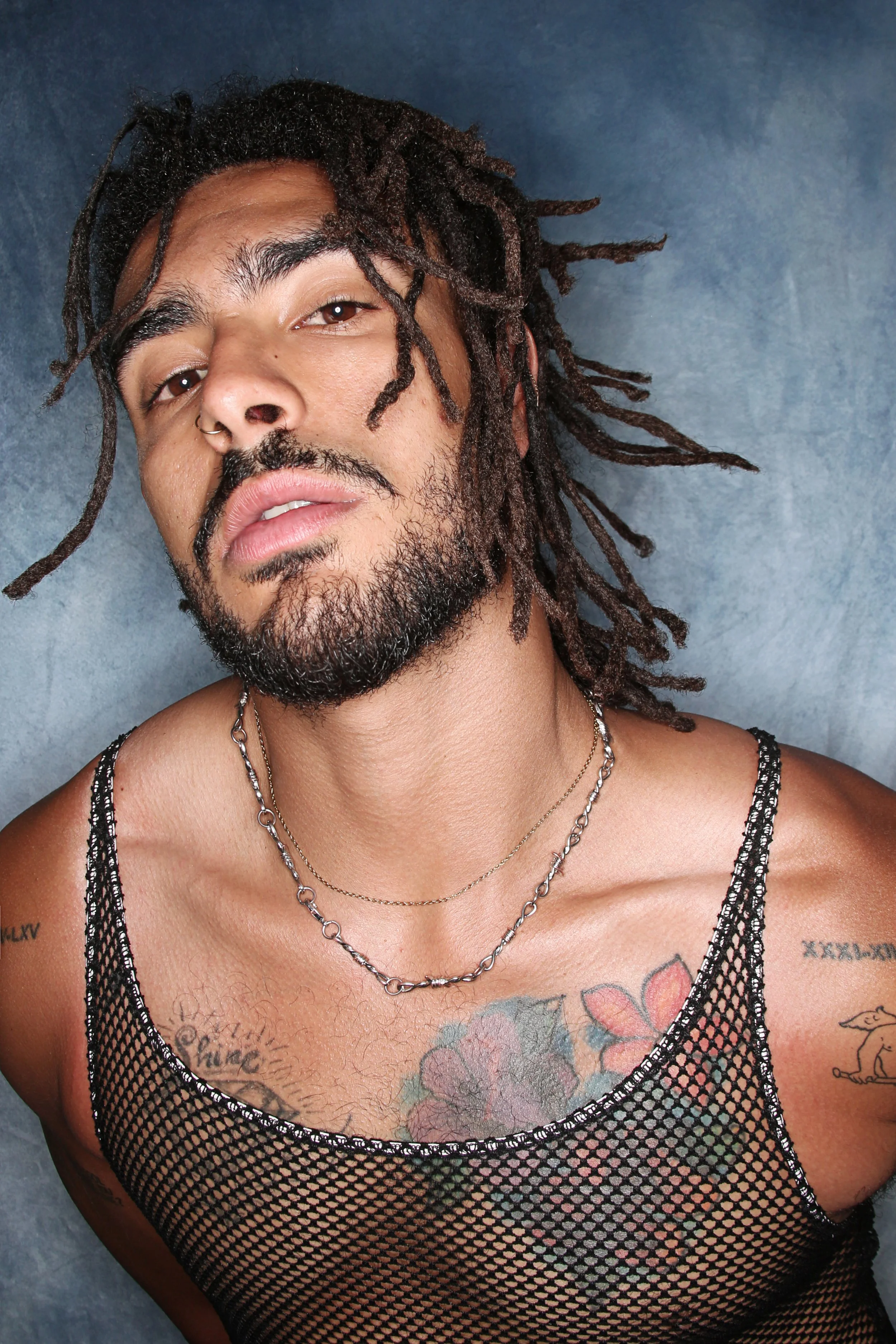 Close-up of a man with dreadlocks and tattoos, wearing a mesh top and necklaces, against a blue background.