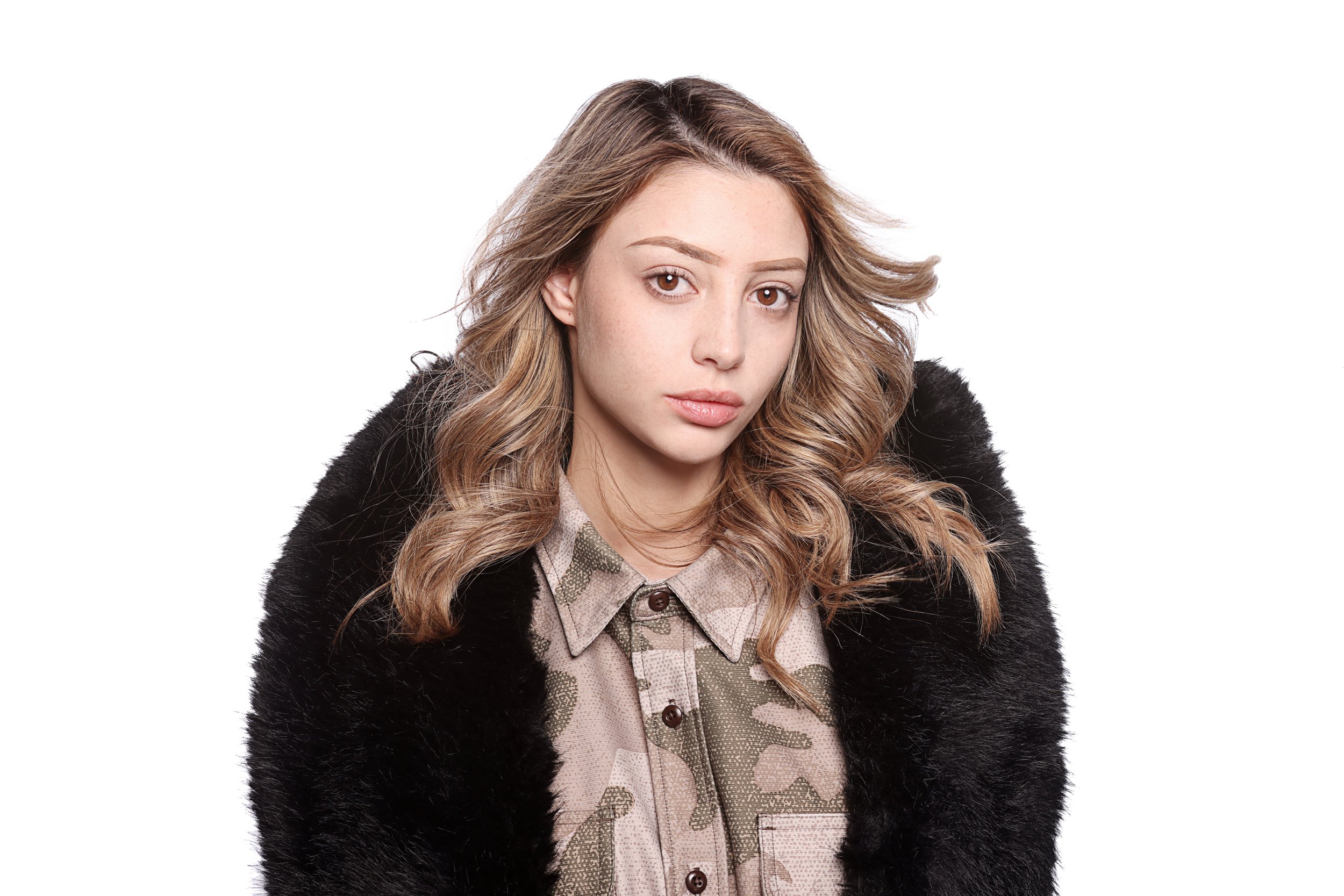 A young woman with wavy, light brown hair and brown eyes wearing a patterned shirt and a black furry jacket, standing against a plain white background.