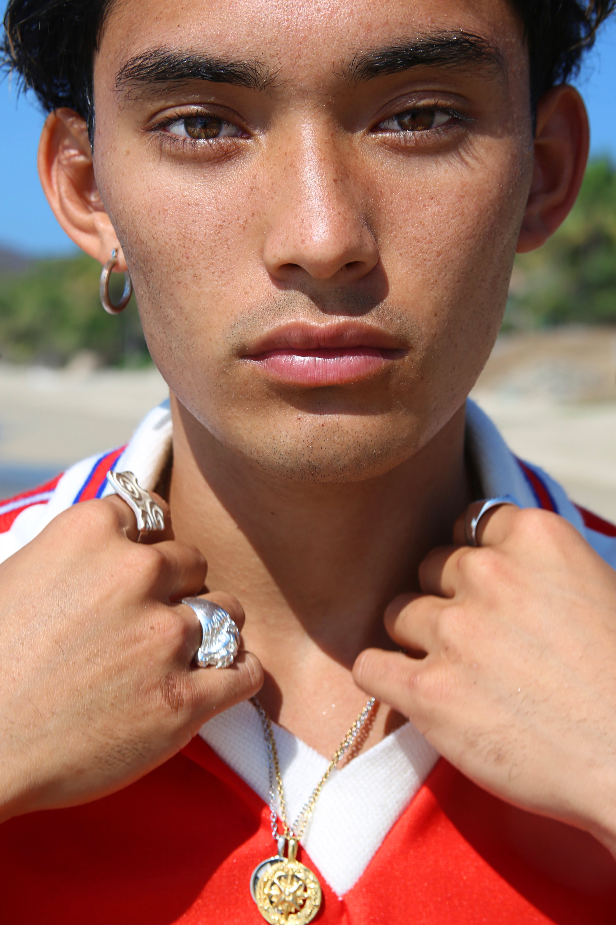 A close-up of a young woman with tan skin, short curly hair, and brown eyes, wearing jewelry including rings, earrings, and a gold pendant. She is holding her necklace with both hands and looking directly at the camera, standing outdoors on a sunny d