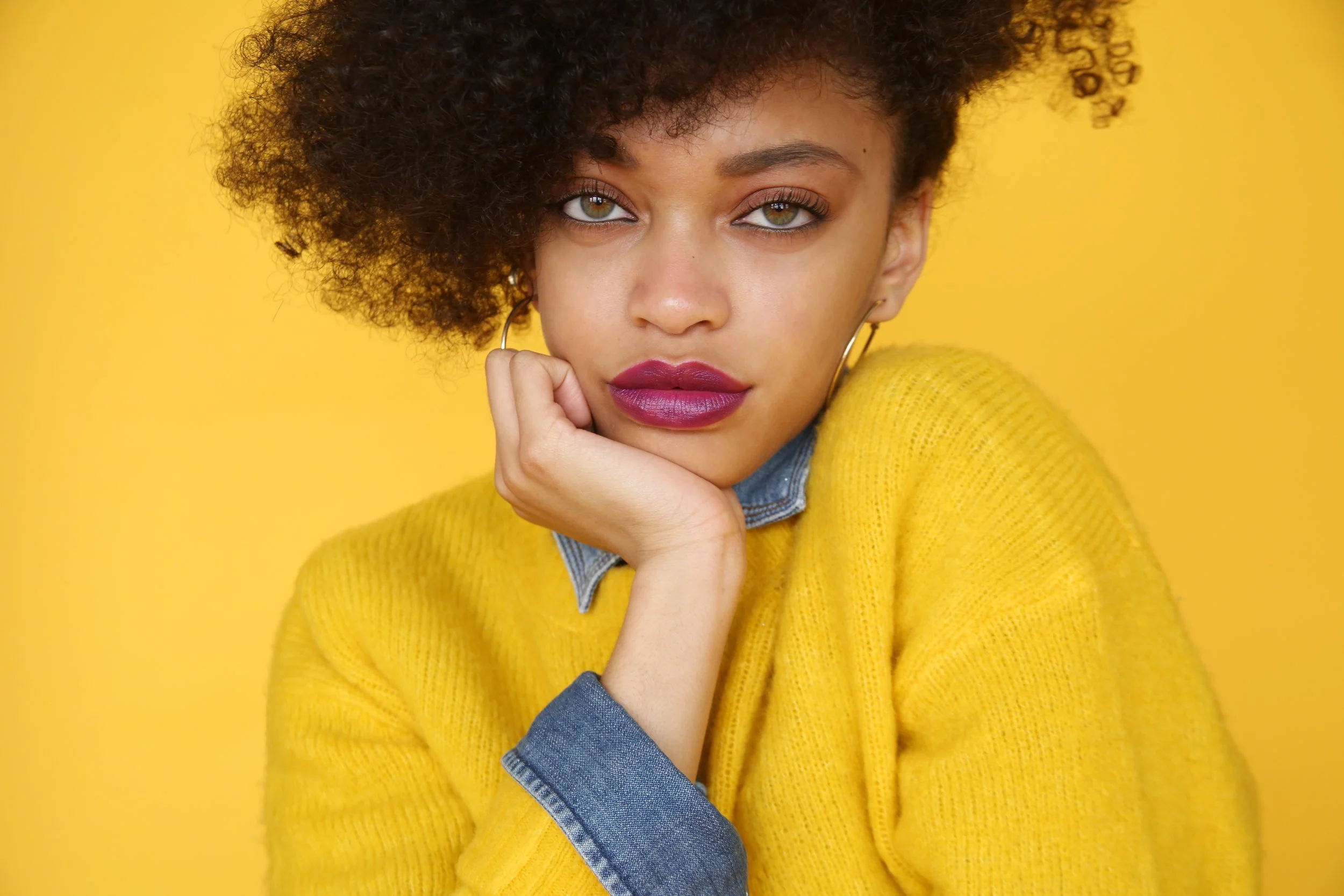 Close-up of a woman with curly hair, wearing a yellow sweater and denim shirt, resting her chin on her hand against a yellow background.