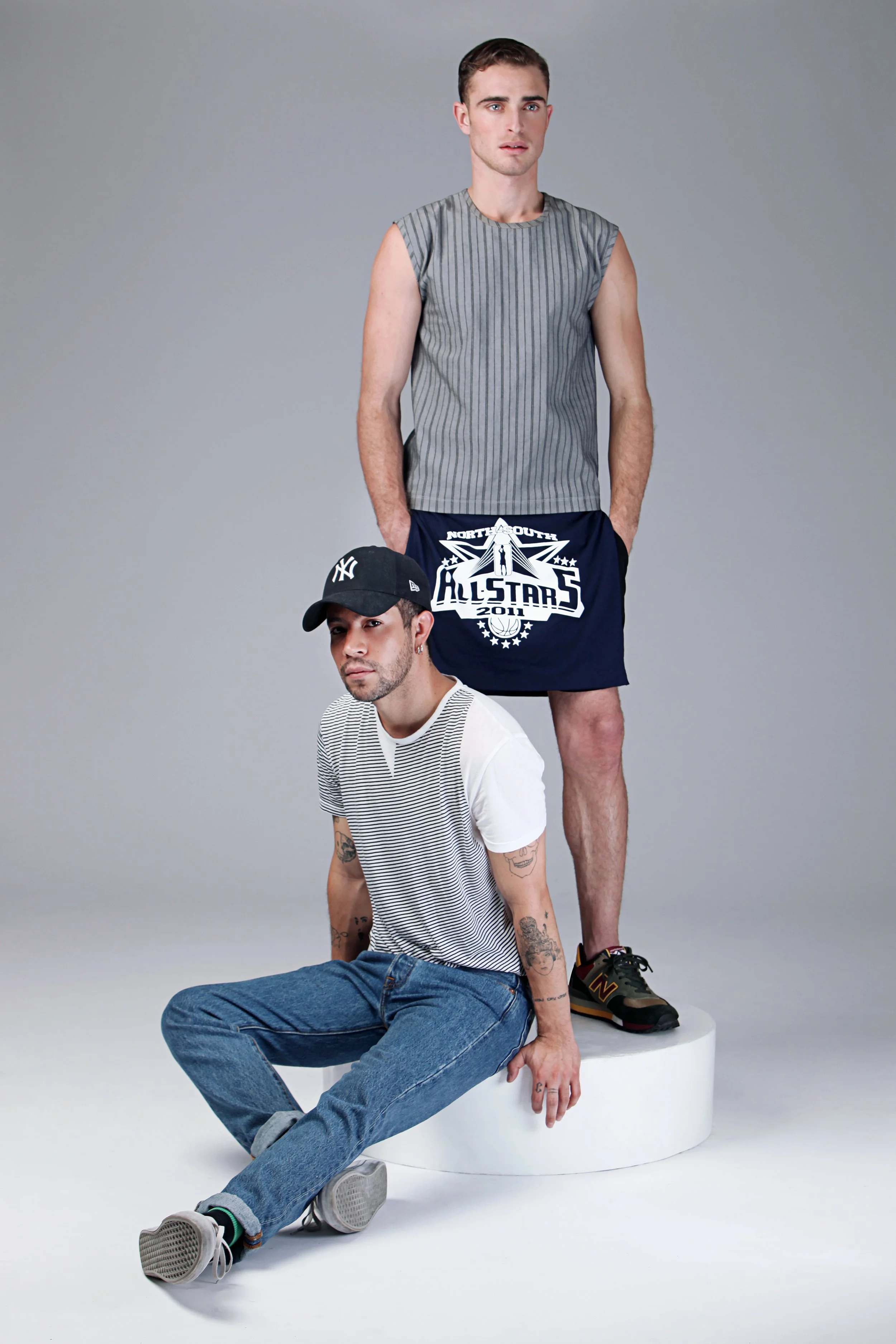 Two young men styled in modern casual fashion, one standing and one sitting on a white circular platform, against a plain light gray background. The standing man wears a sleeveless gray striped shirt and navy shorts with a sports logo, while the seat