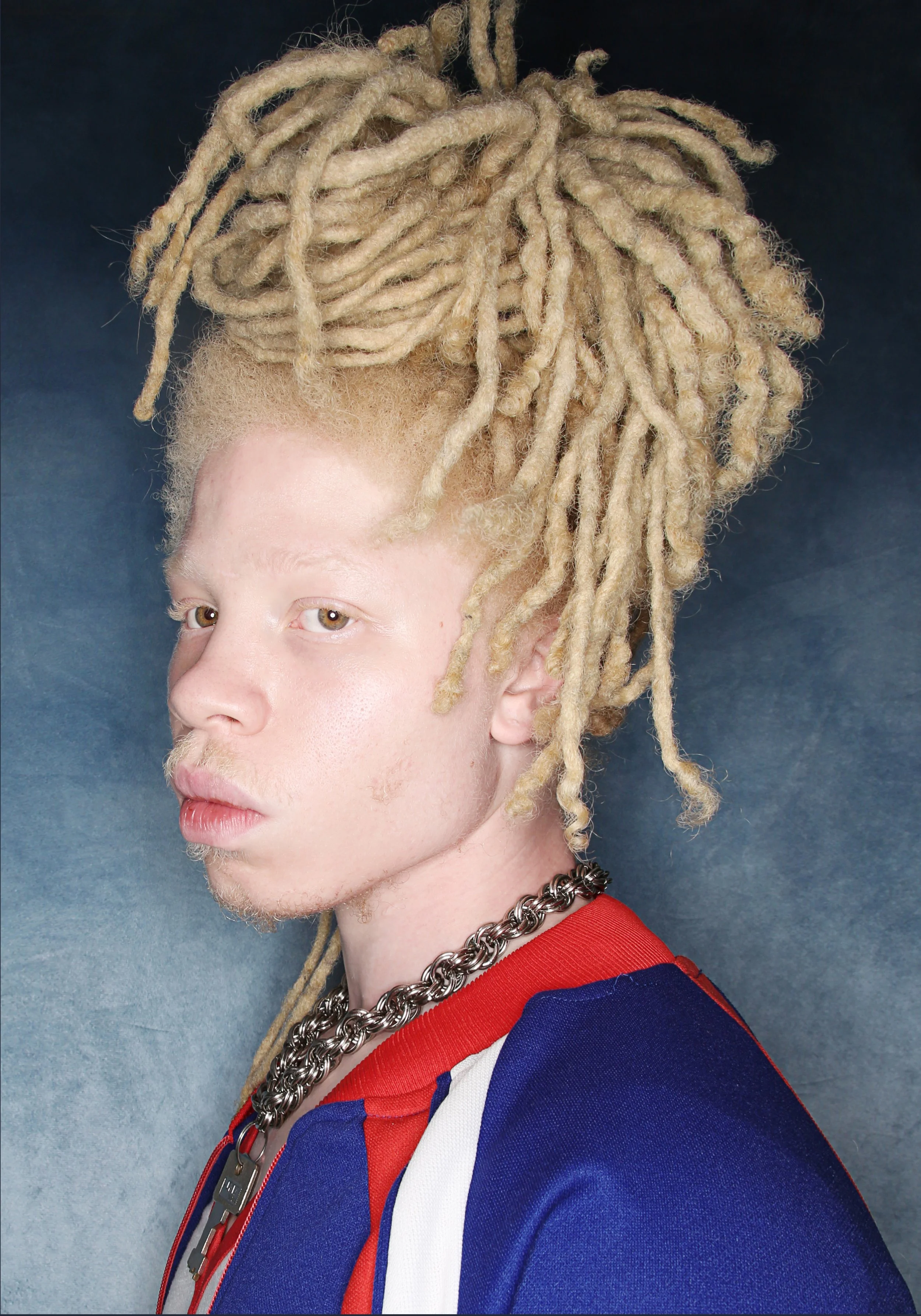 Young man with blond dreadlocks wearing a red, white, and blue sports jacket, silver chain necklace, and a silver dog tag pendant, posing against a dark background.