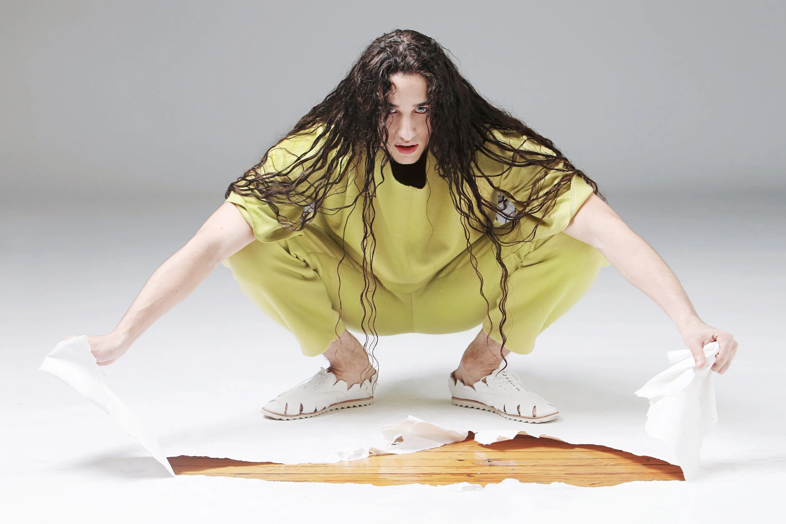 A woman with long, curly black hair wearing a yellow jumpsuit, crouched down with her hands on the floor, looking intently at the camera. The background is plain gray.