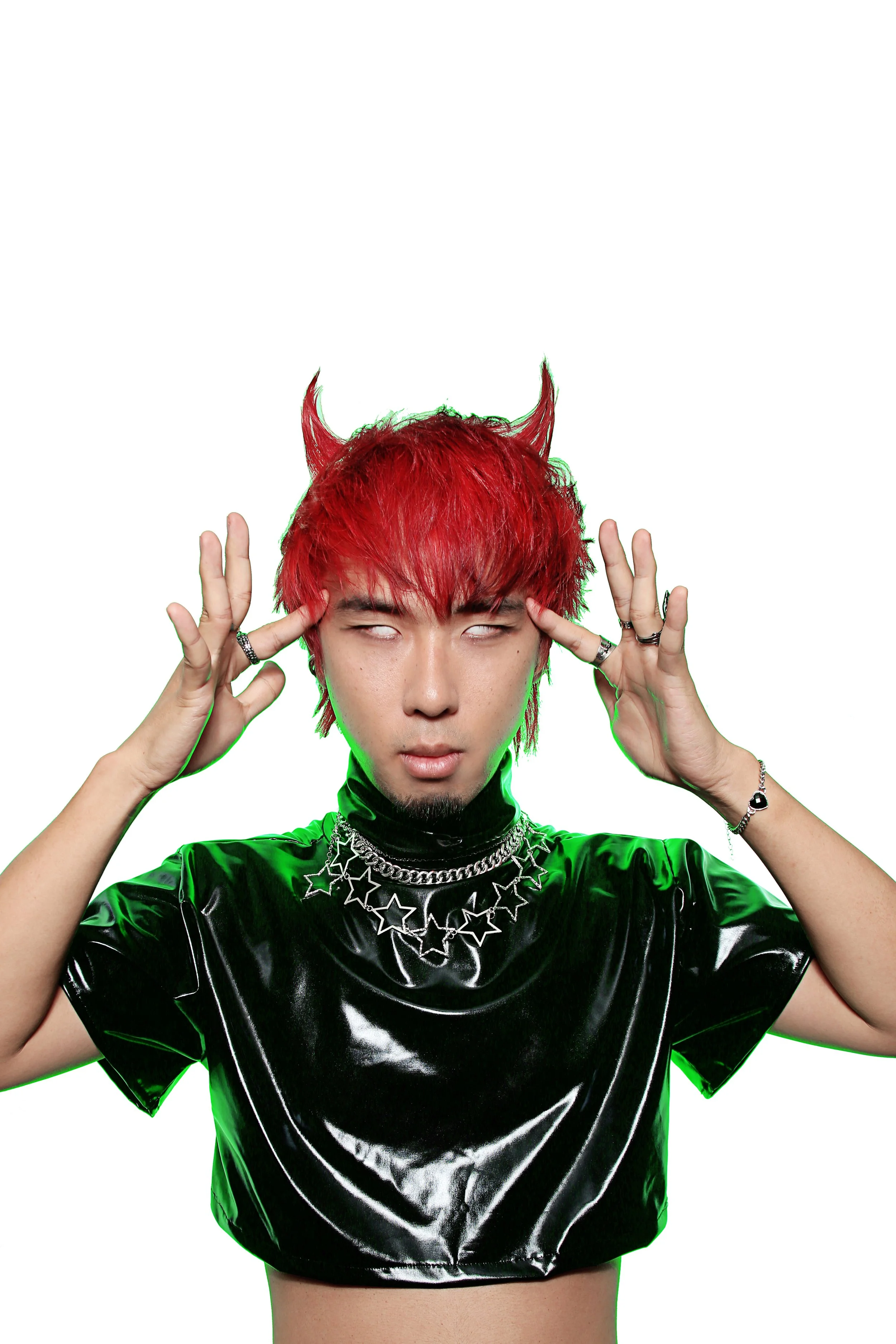 Person with red hair styled into devil horns, wearing a shiny black top and star jewelry, tilting their head with eyes closed, holding fingers near temples, against a white background.