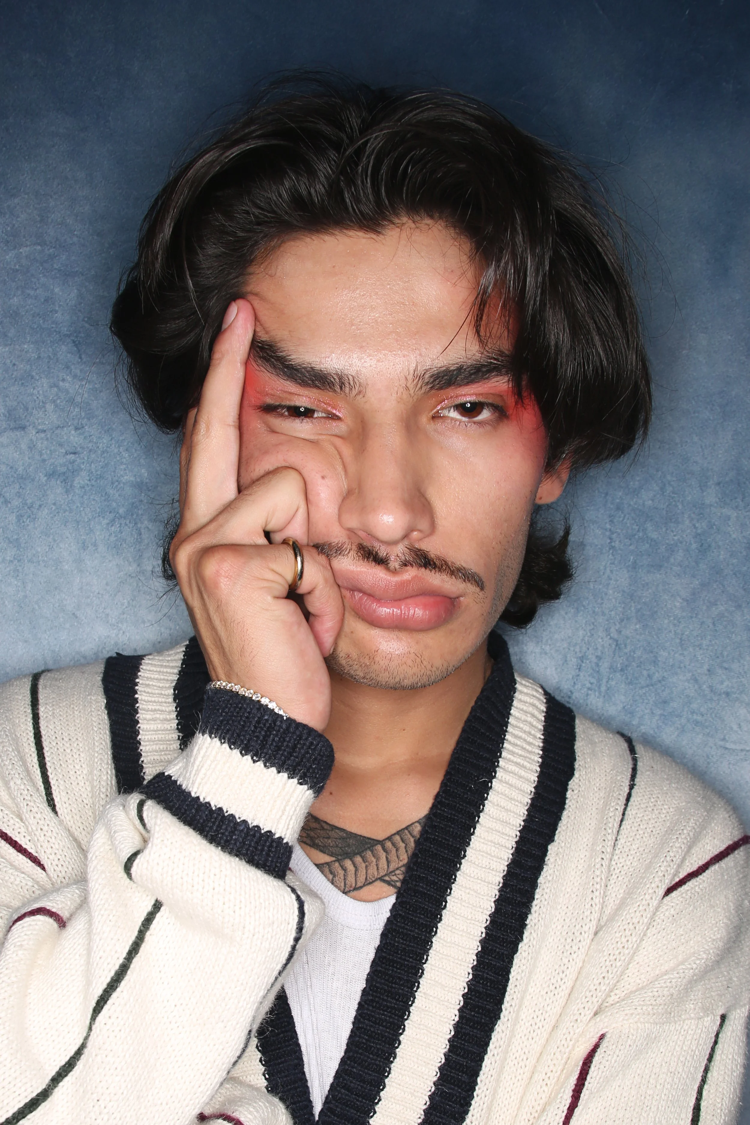 A young man with dark, wavy hair and facial hair making a peace sign near his eye. He is wearing a cream-colored knit sweater with black and red stripes, and is partially covering his face with his hand. The background is a textured bluish surface.