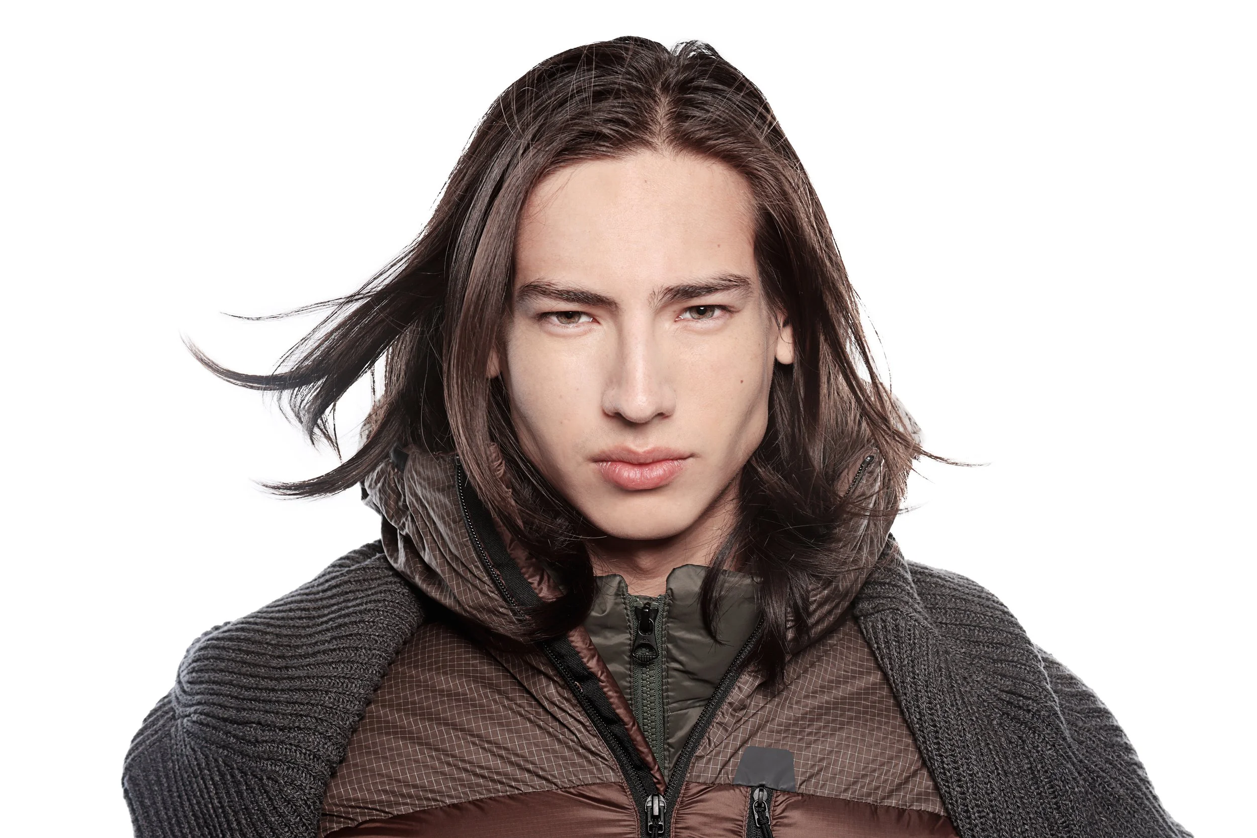 Portrait of a young woman with long dark hair wearing a brown and black jacket, looking directly at the camera against a white background.