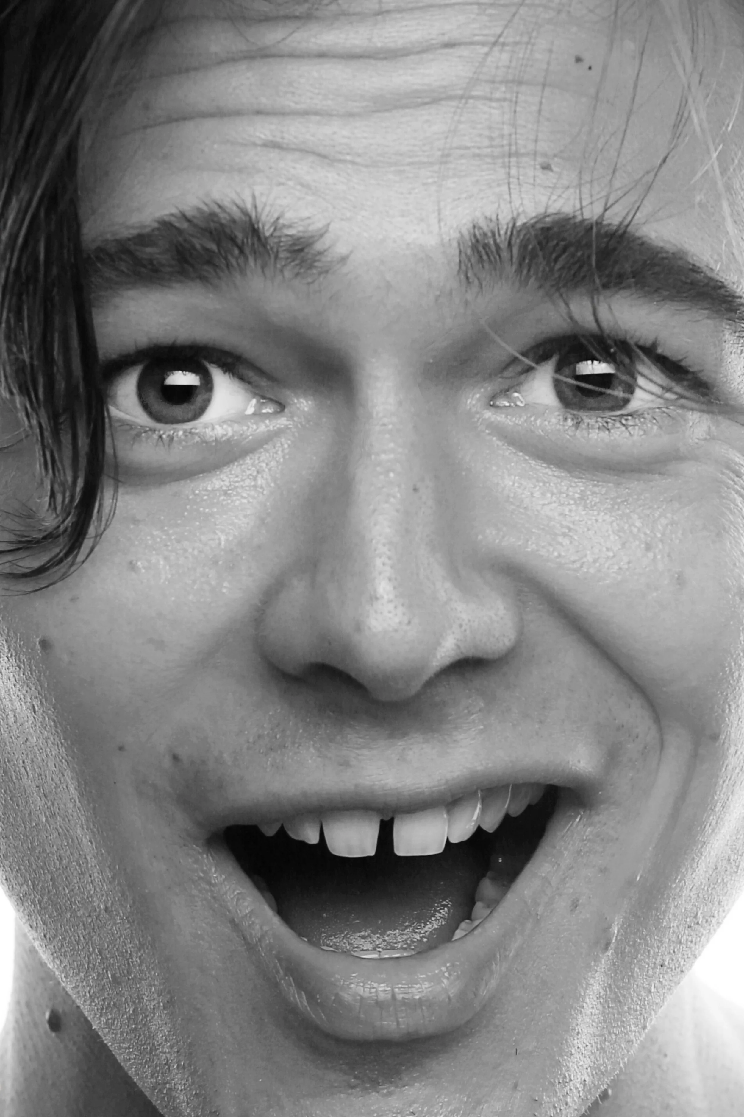 A close-up black and white photo of a person's face showing only the eyes, nose, and mouth, with an open-mouthed smile showing teeth.