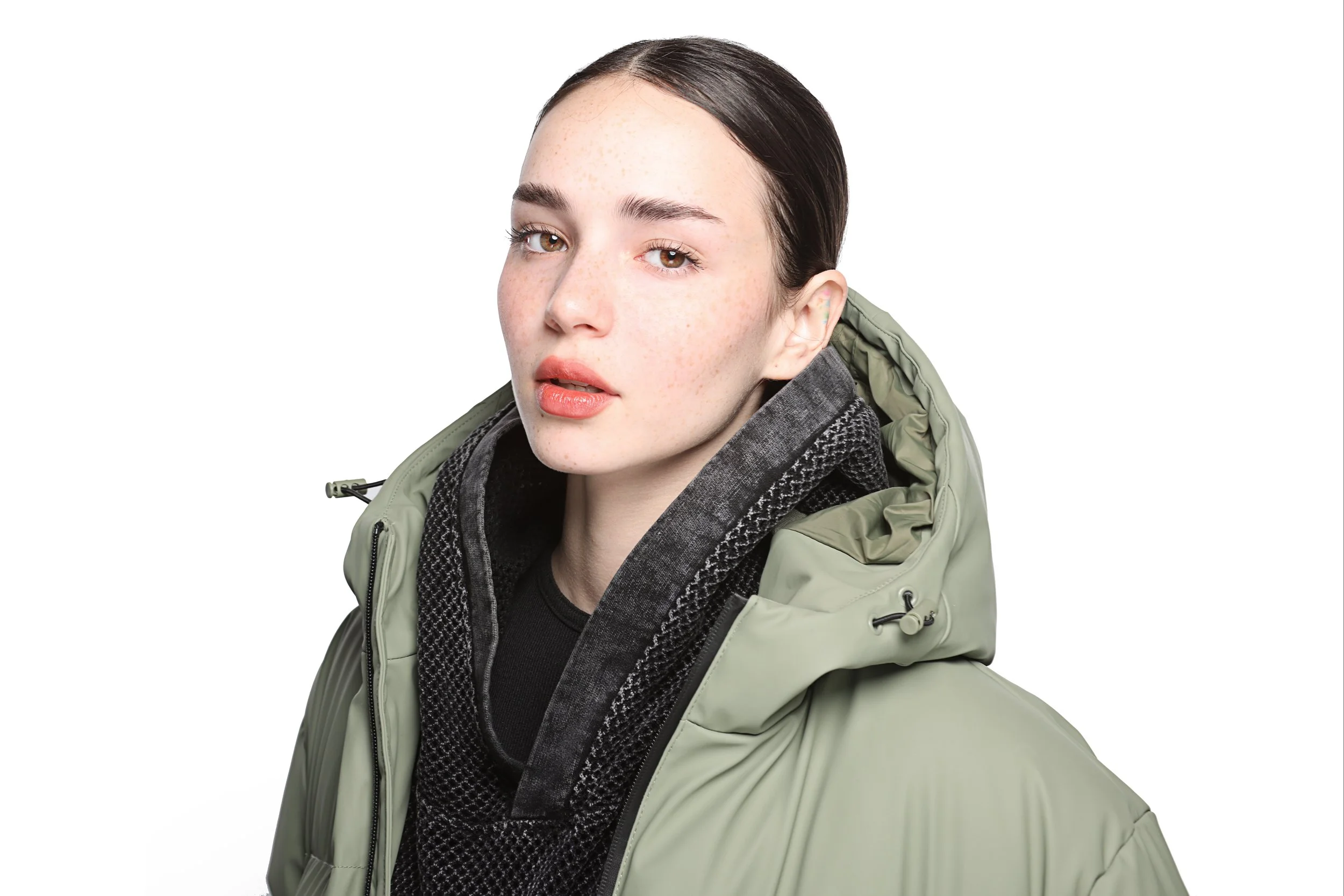 A young woman with brown hair, light skin, and freckles, wearing a black shirt, a patterned gray sweater, and an olive-green jacket, looking at the camera with a neutral expression.