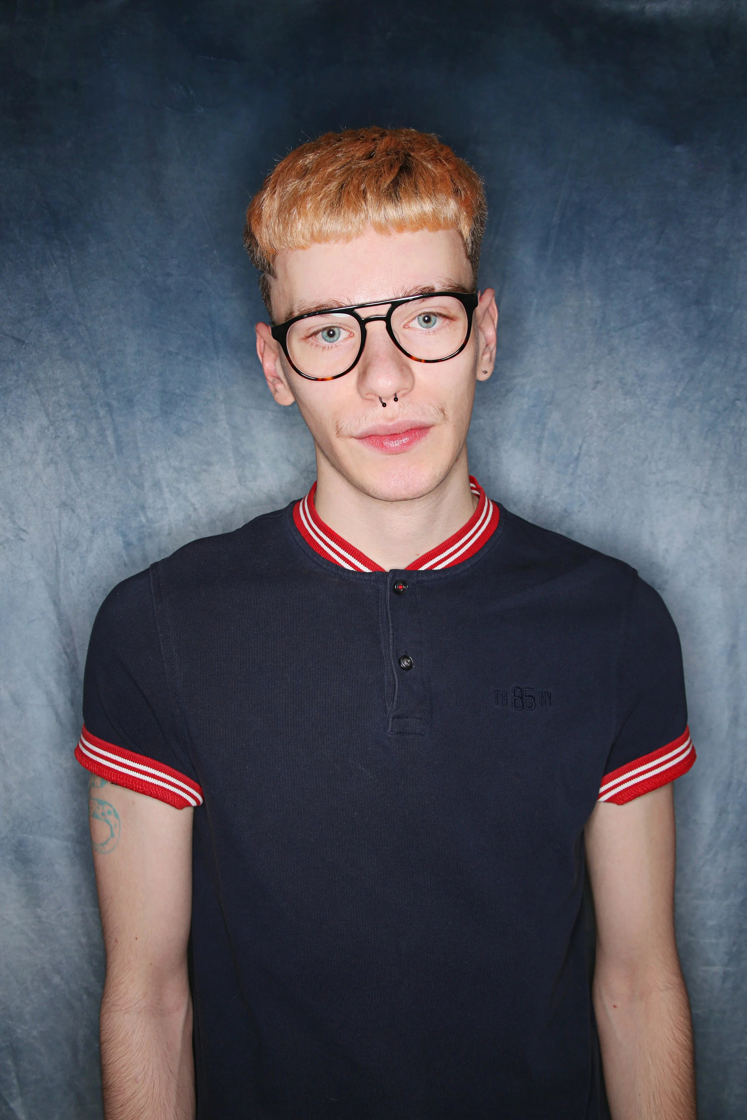 A young man with short red hair and round black glasses, wearing a navy blue shirt with red and white striped trim on the collar and sleeves, standing against a gray background.