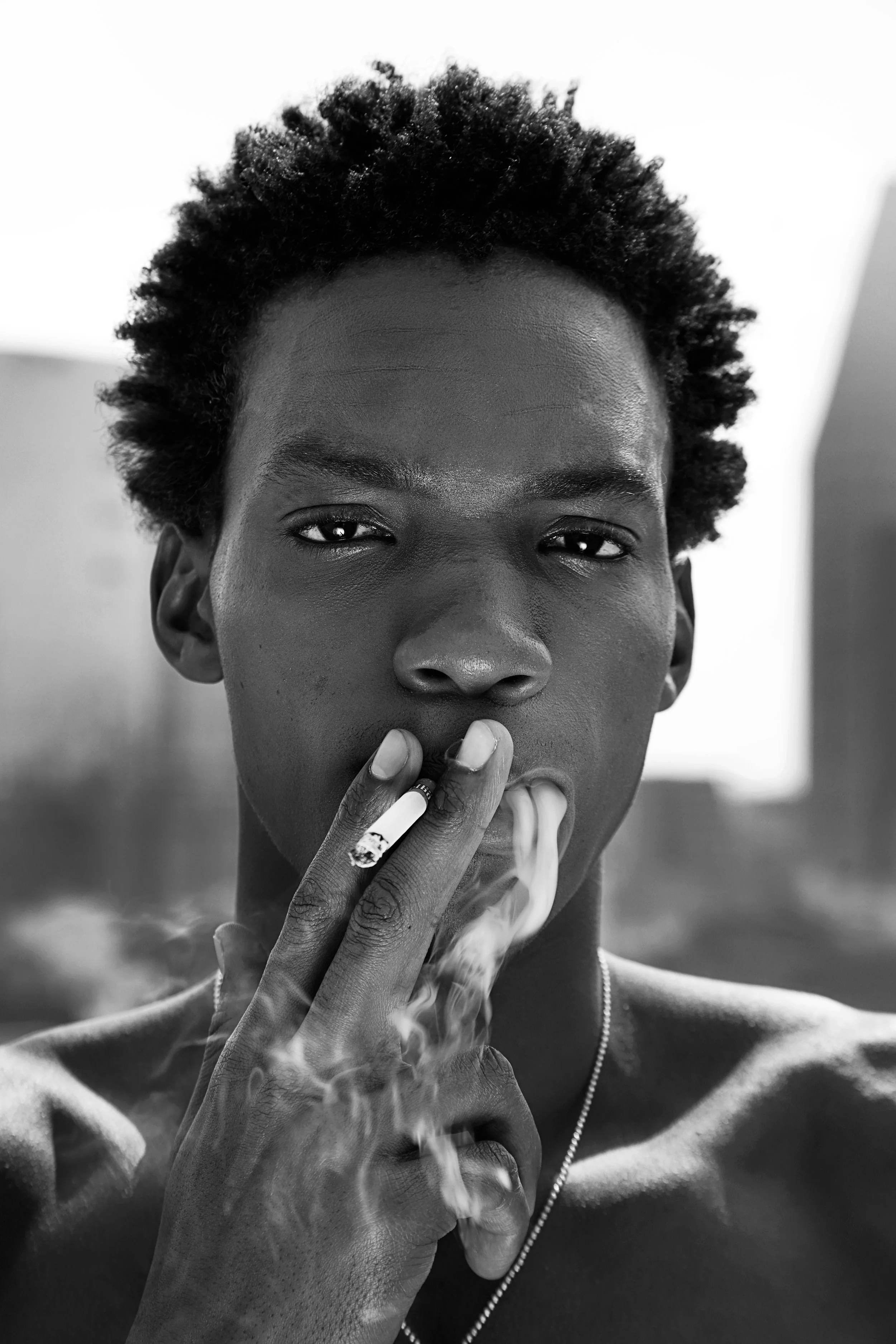 Close-up black and white photograph of a person with short curly hair smoking, holding a cigarette with fingers, with smoke rising, and a blurred background.