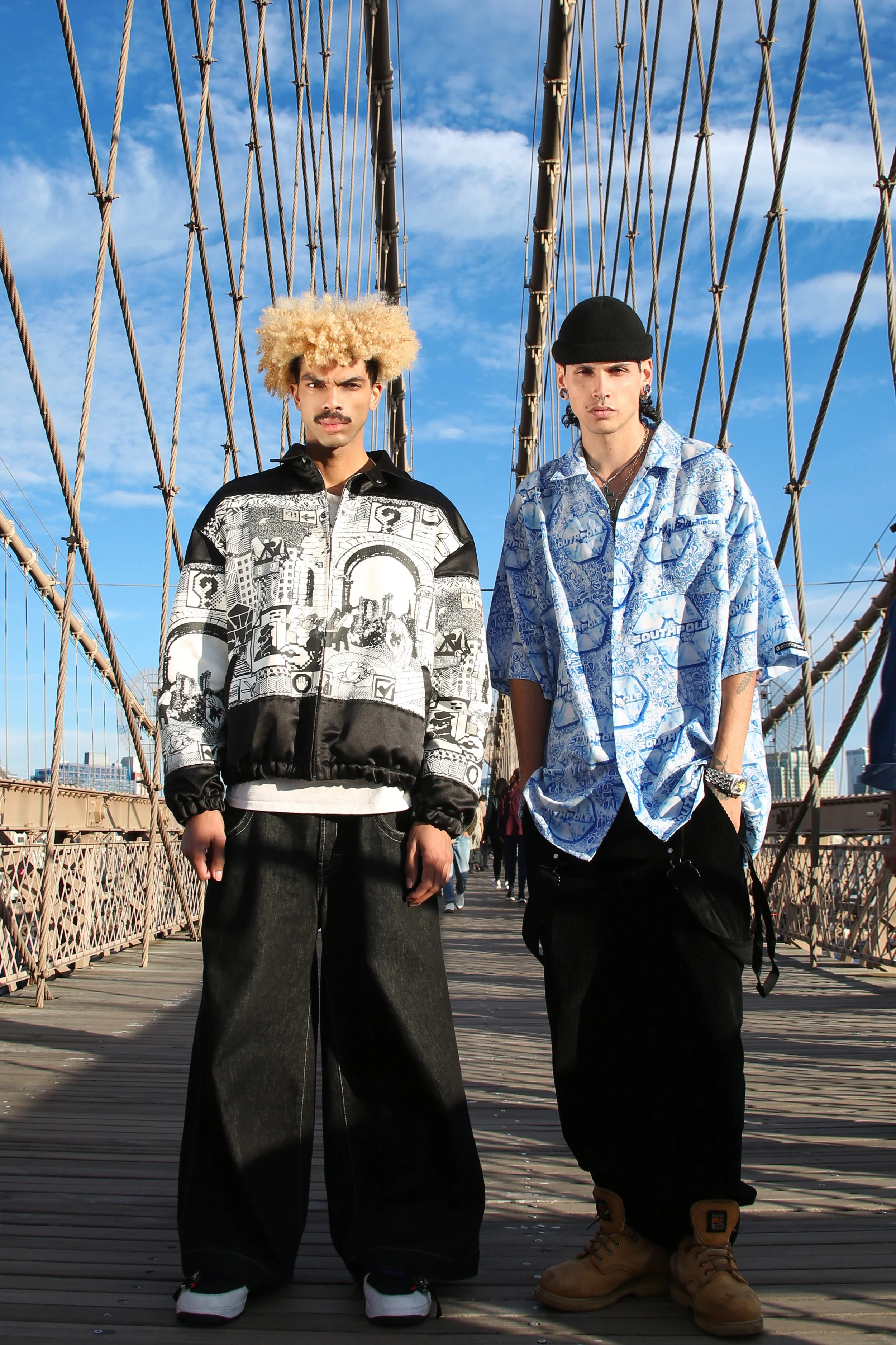 Two young men standing on the Brooklyn Bridge in New York City with a blue sky and clouds in the background.