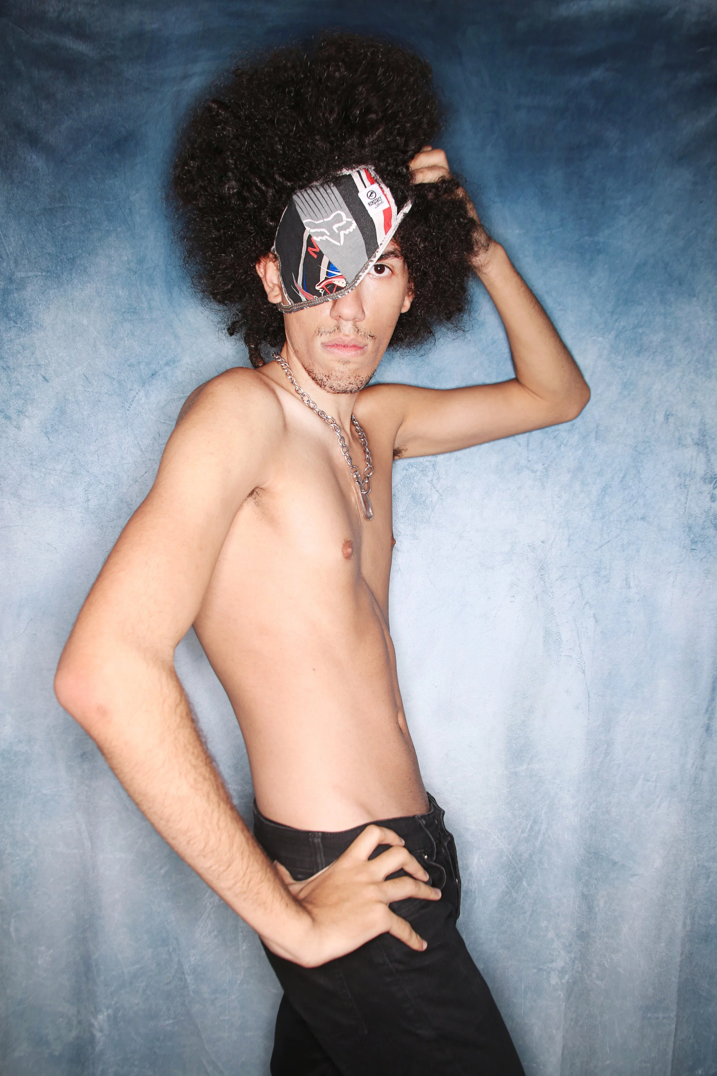 A shirtless young man with curly hair and a chain necklace, wearing a face mask over one eye and black jeans, posing against a textured blue wall.