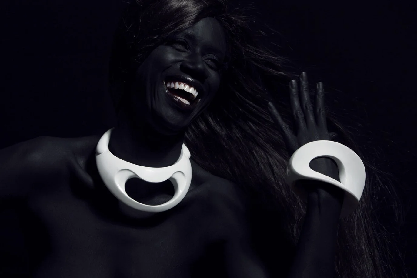 A Black woman is smiling and laughing while wearing a white necklace and bracelet with large loop designs. Her hair is flowing and she is in a dark setting.