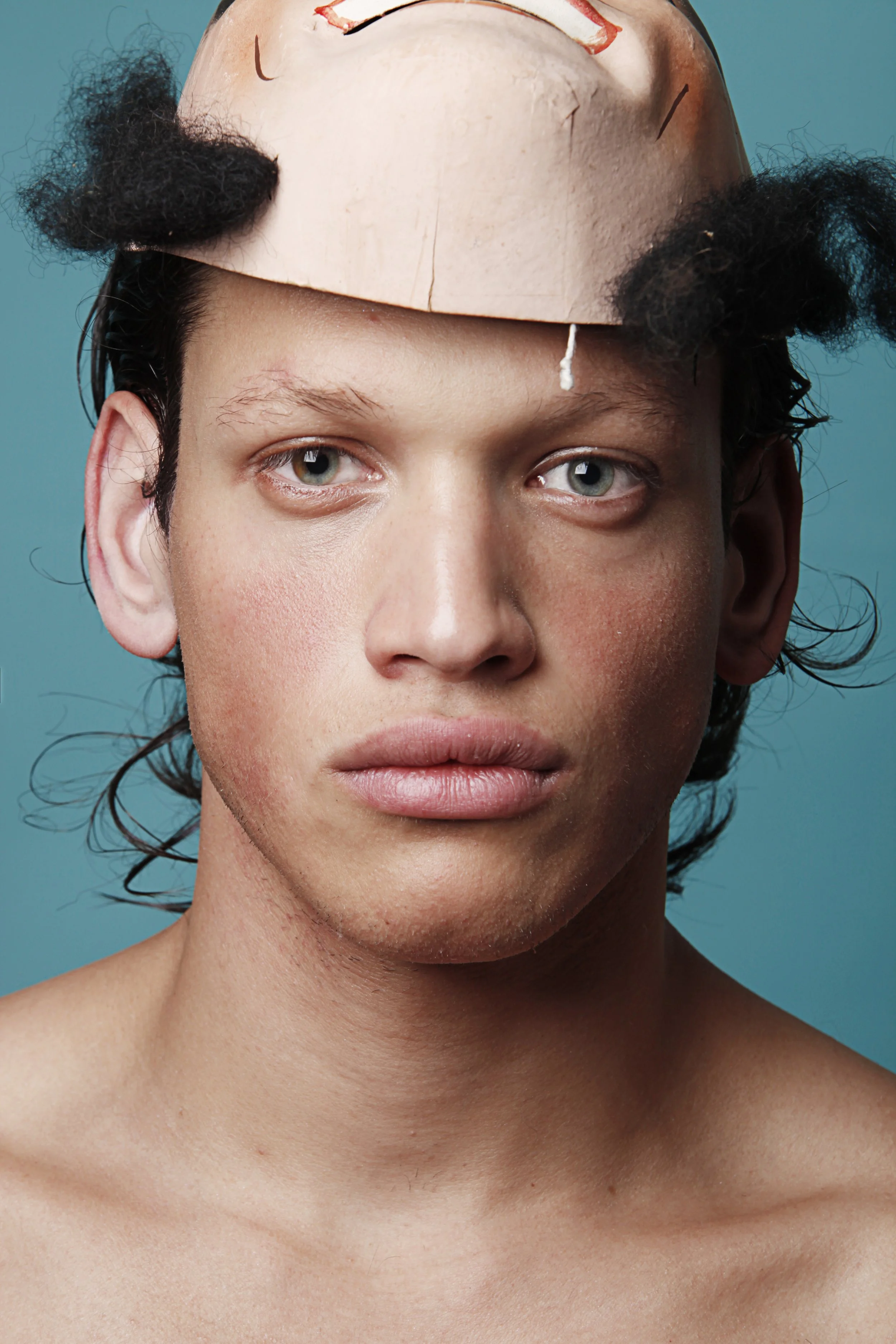 A person with short dark curly hair wearing a paper mask on their forehead and no shirt, staring directly at the camera against a teal background.
