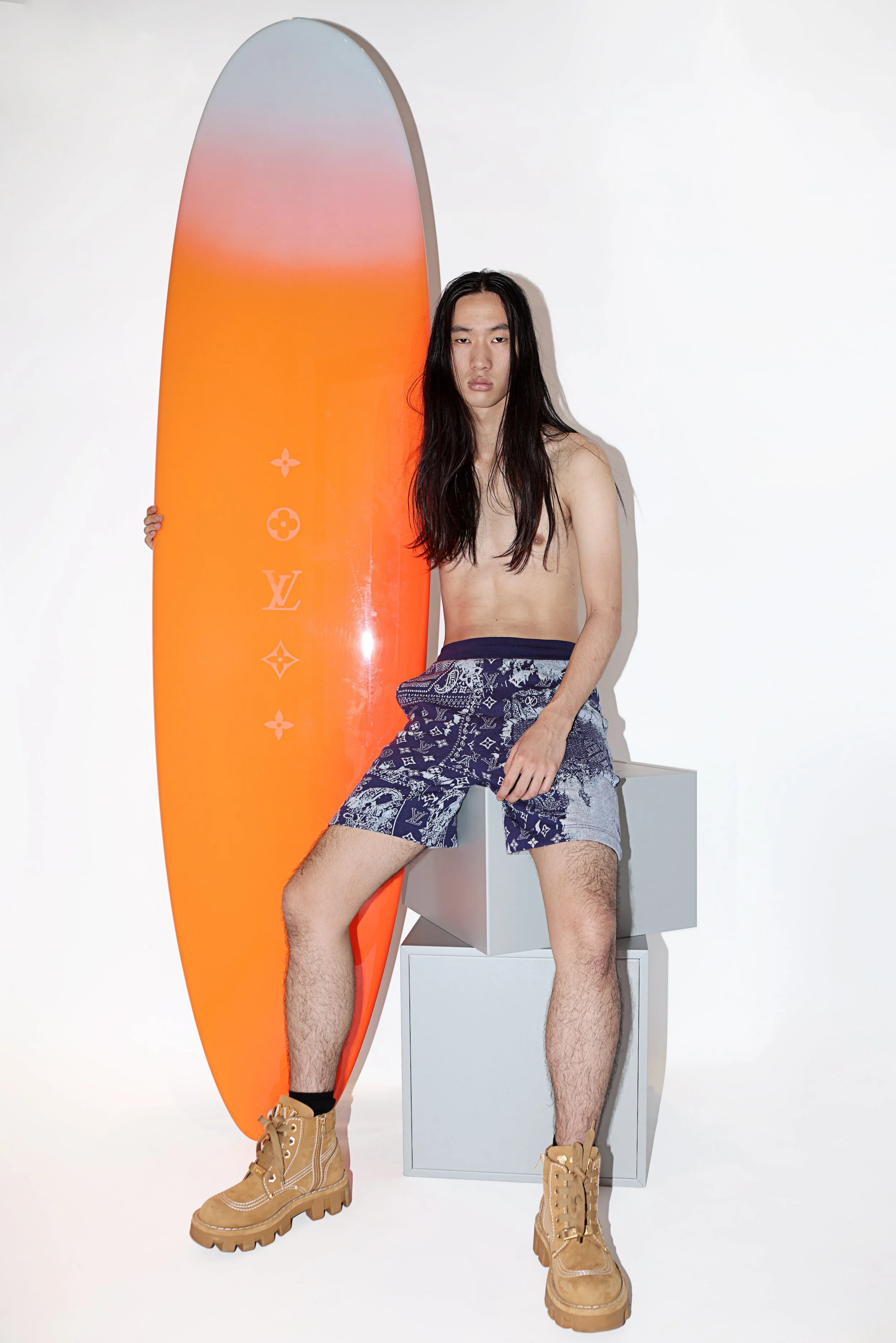 A young man with long black hair is standing next to an orange surfboard with Louis Vuitton logos. He is shirtless, wearing patterned shorts, tan boots, and black socks, with one hand resting on a white block and the other holding the surfboard.