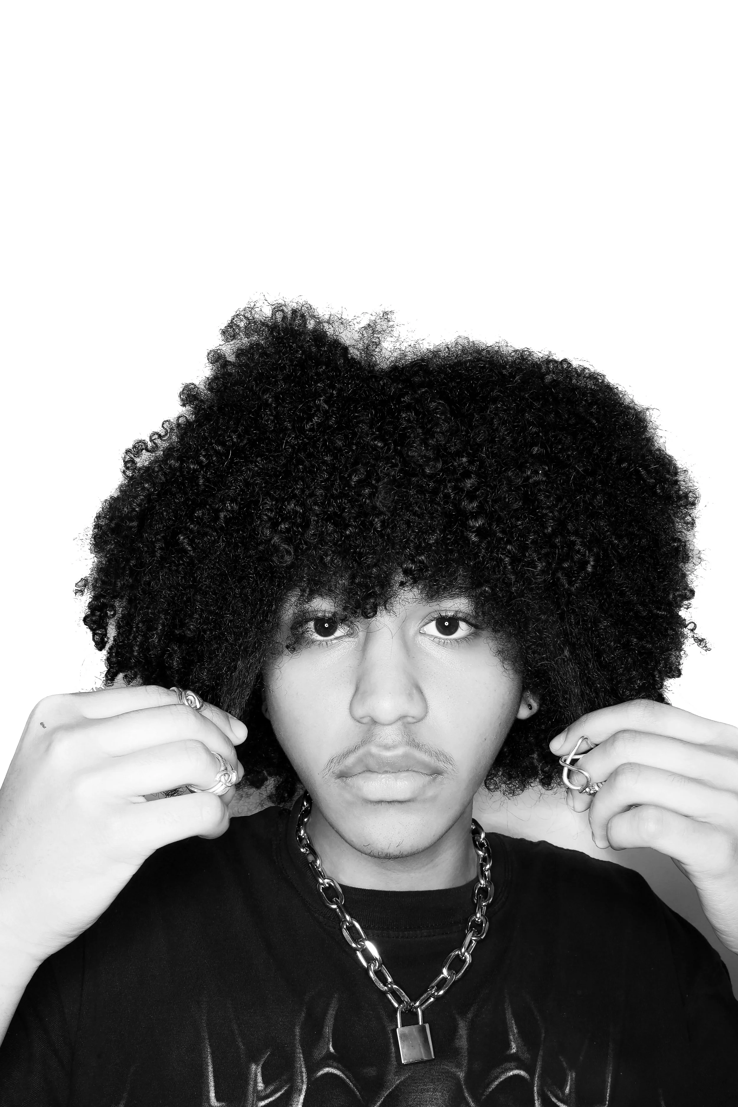 A black-and-white close-up photo of a man with curly hair and multiple rings adjusting his hair, wearing a chain necklace and a T-shirt.