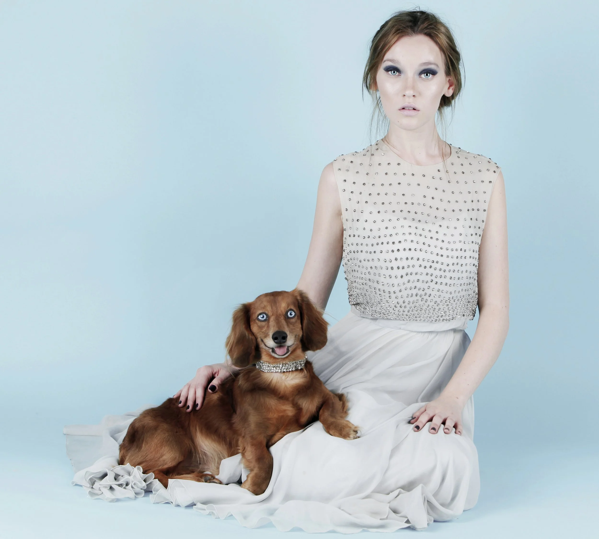 A young woman with short, wavy, reddish-brown hair and makeup, wearing a sleeveless beige top with metallic embellishments, sitting on a white fabric backdrop with a light blue background, holding a brown dachshund with blue eyes and a sparkly collar