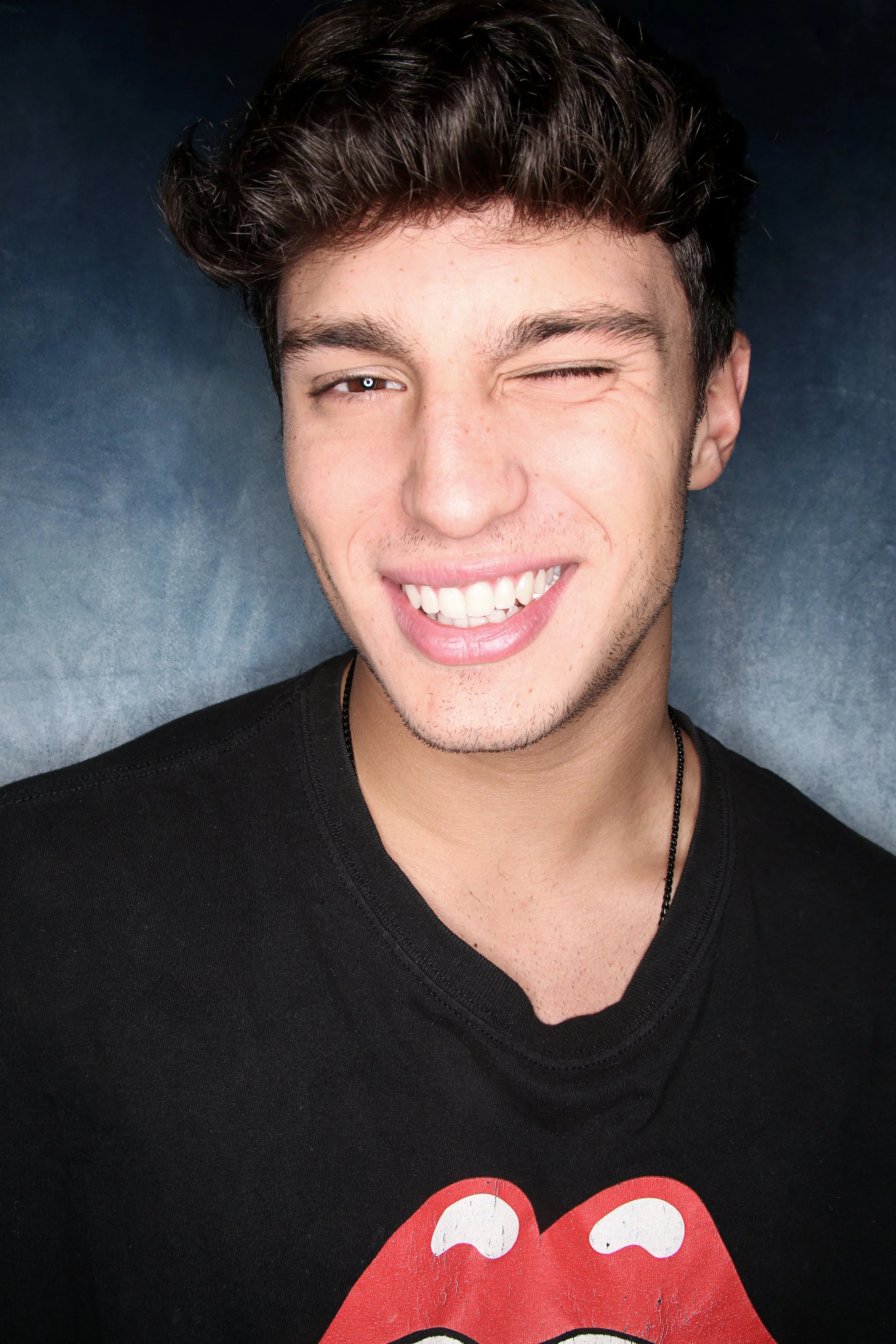 A young man with dark, styled hair winking, smiling, wearing a black T-shirt with a red Rolling Stones tongue logo, standing against a dark gradient background.