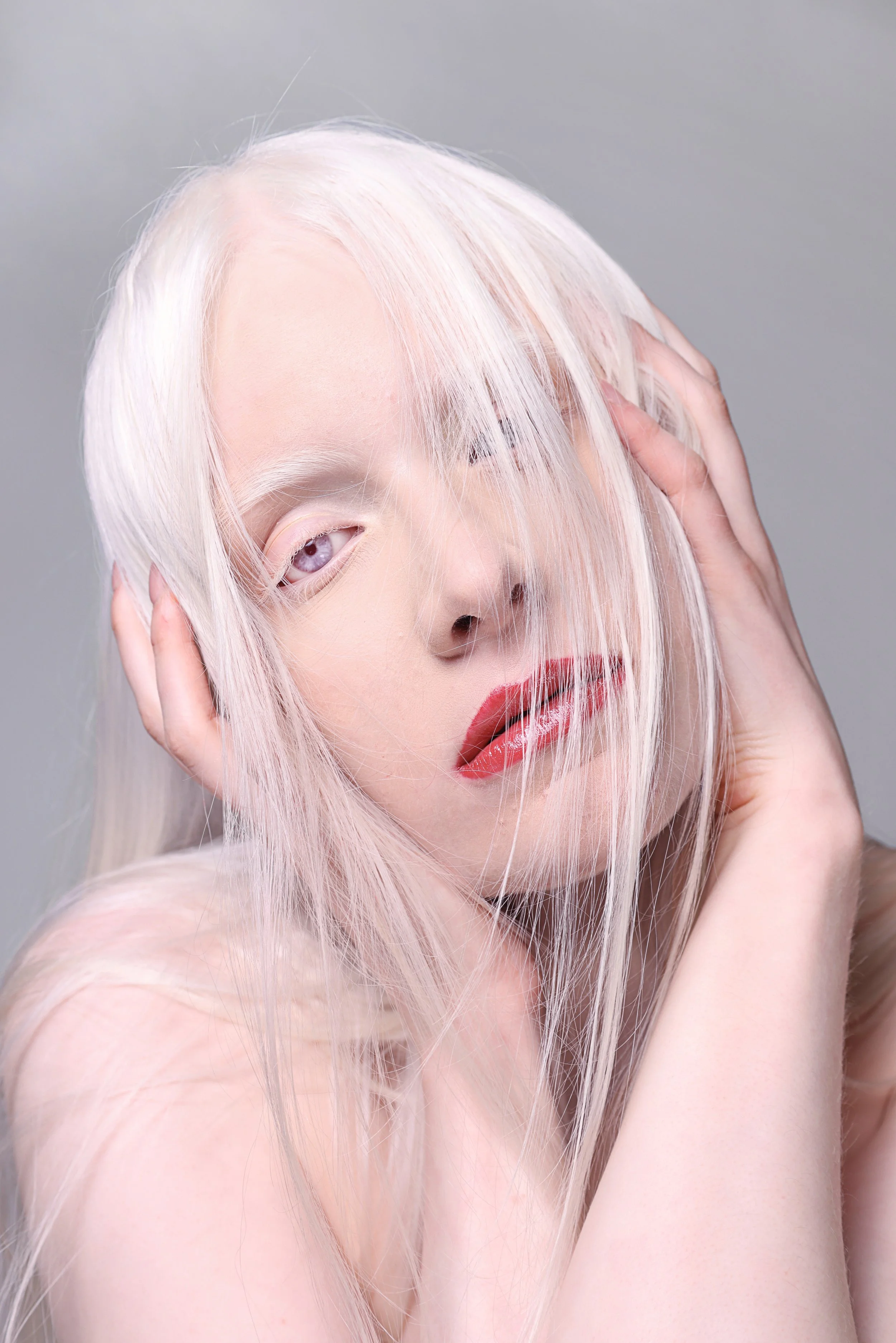 A woman with long, platinum blonde hair covering part of her face, wearing red lipstick, and touching her head with both hands against a gray background.