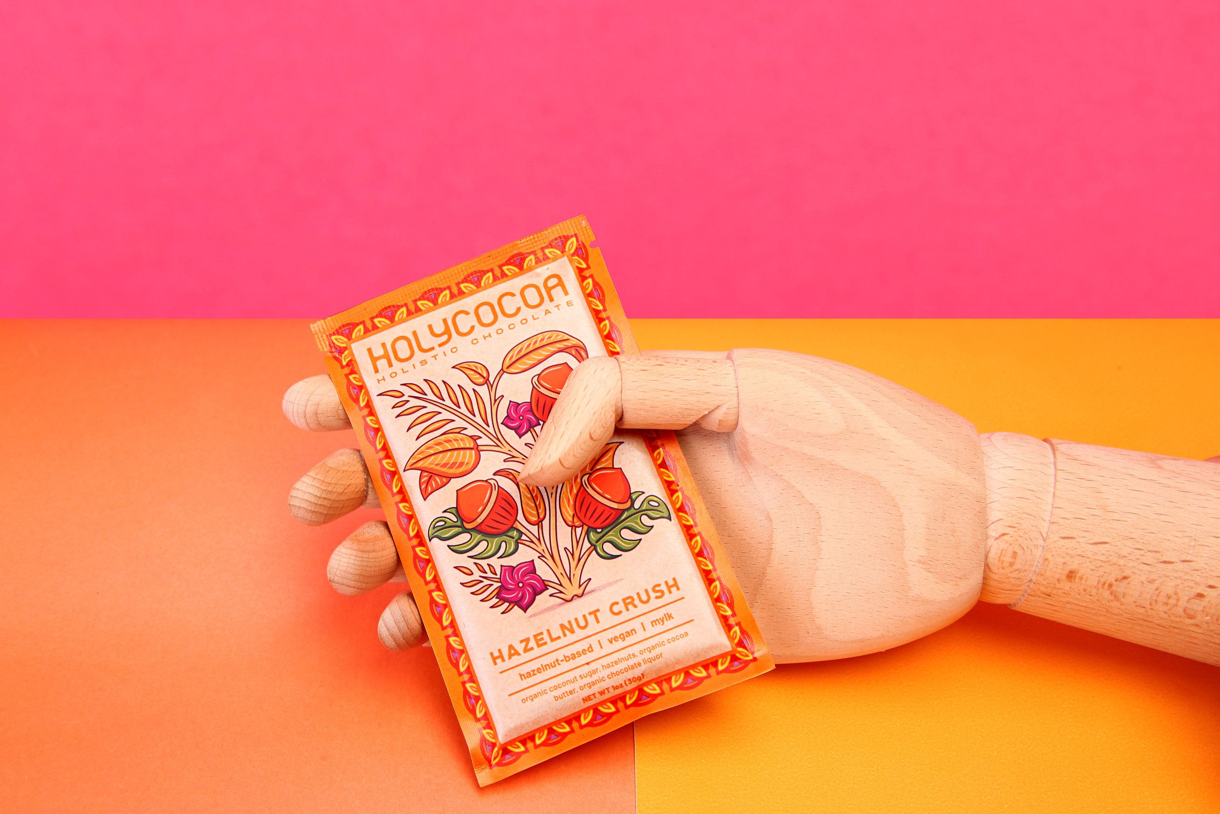 A wooden mannequin hand holding a colorful package of Holy cacao hazelnut crunch vegan chocolate against a vibrant pink and orange background.