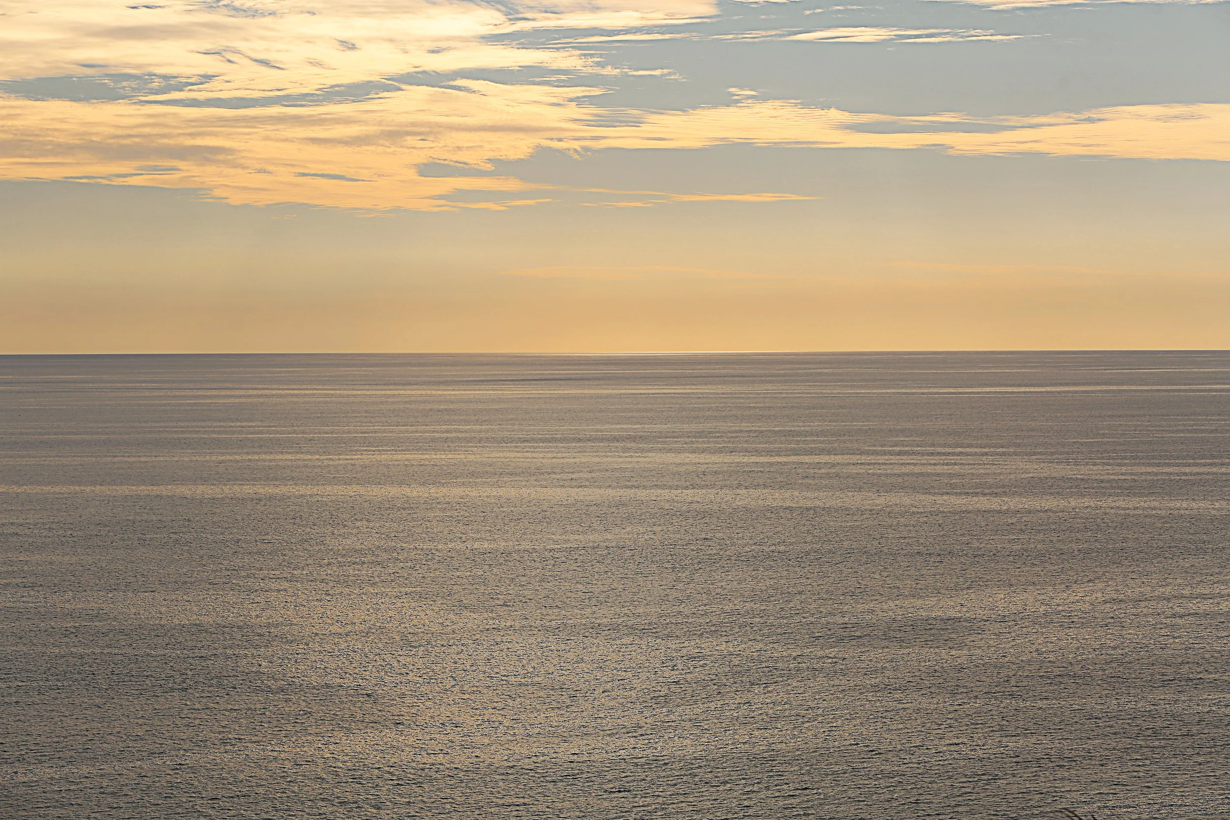 Sunset over a calm ocean with light-colored clouds in the sky.
